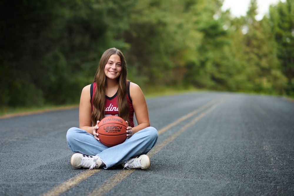 Smiling teen girl sitting cross-legged on a road holding a basketball for her senior portrait photos.
