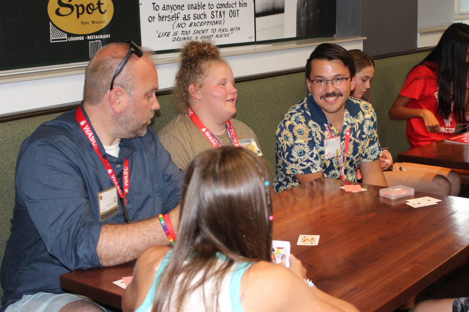 Three adults smile and engage in a card game together at a wooden table during a campus social event.