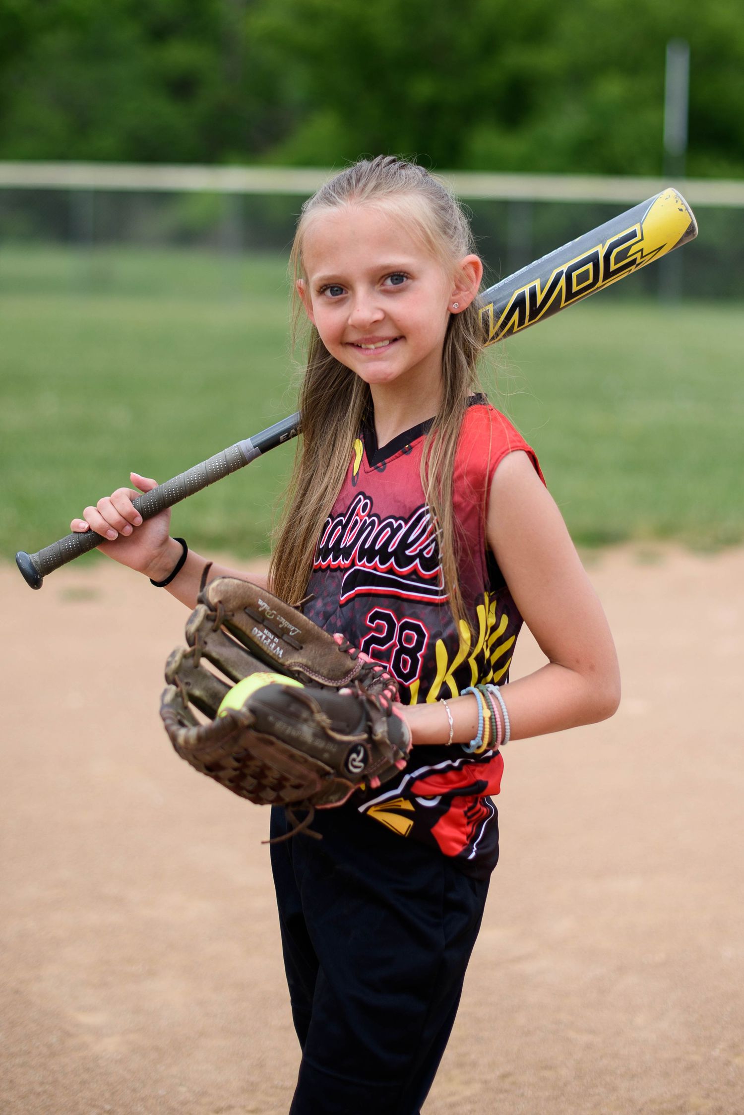 Girl in Cardinals #28 softball jersey posing with black bat and worn glove on dirt field.