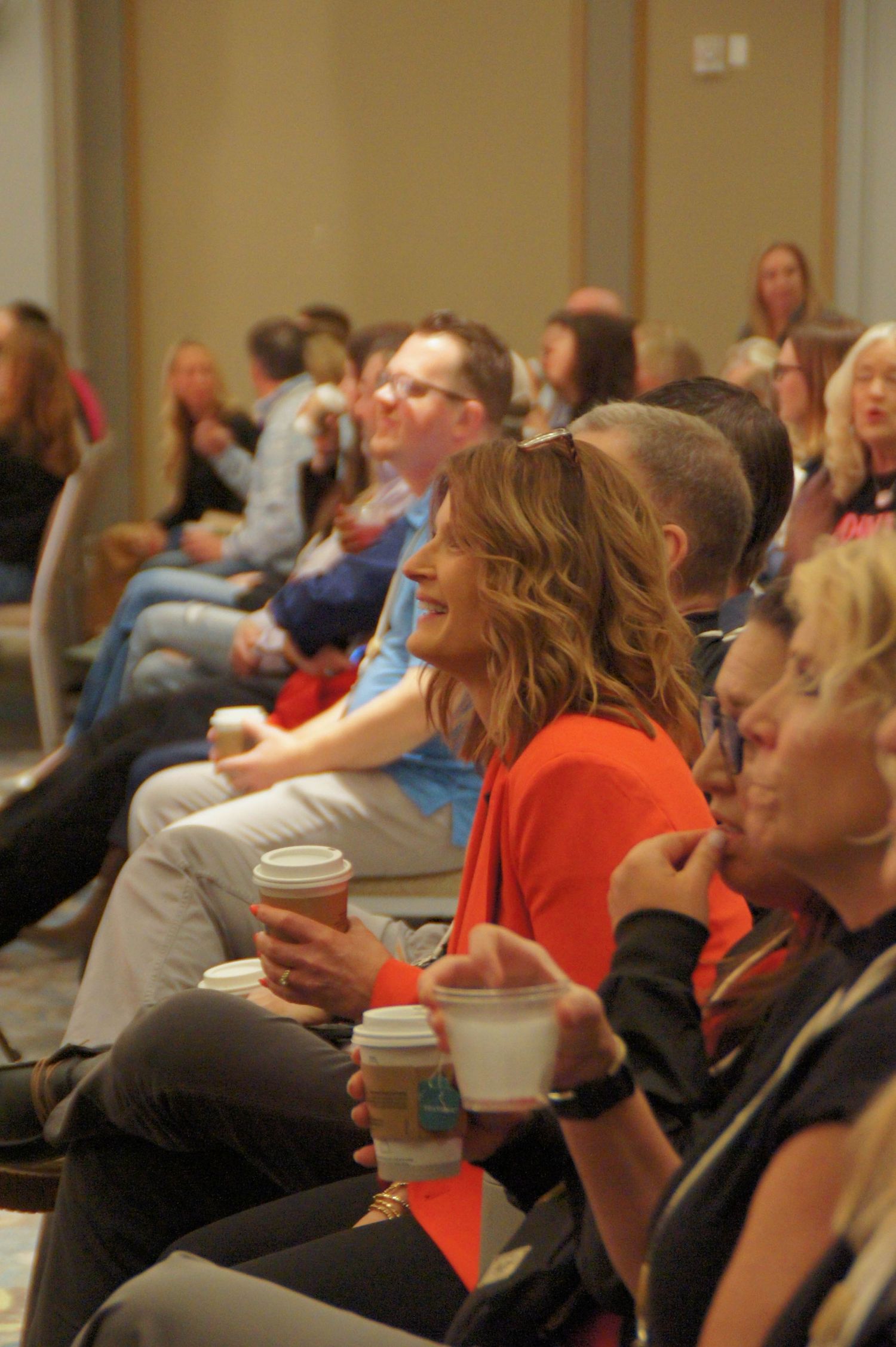 Audience members smile and hold coffee cups at an indoor conference or seminar event.