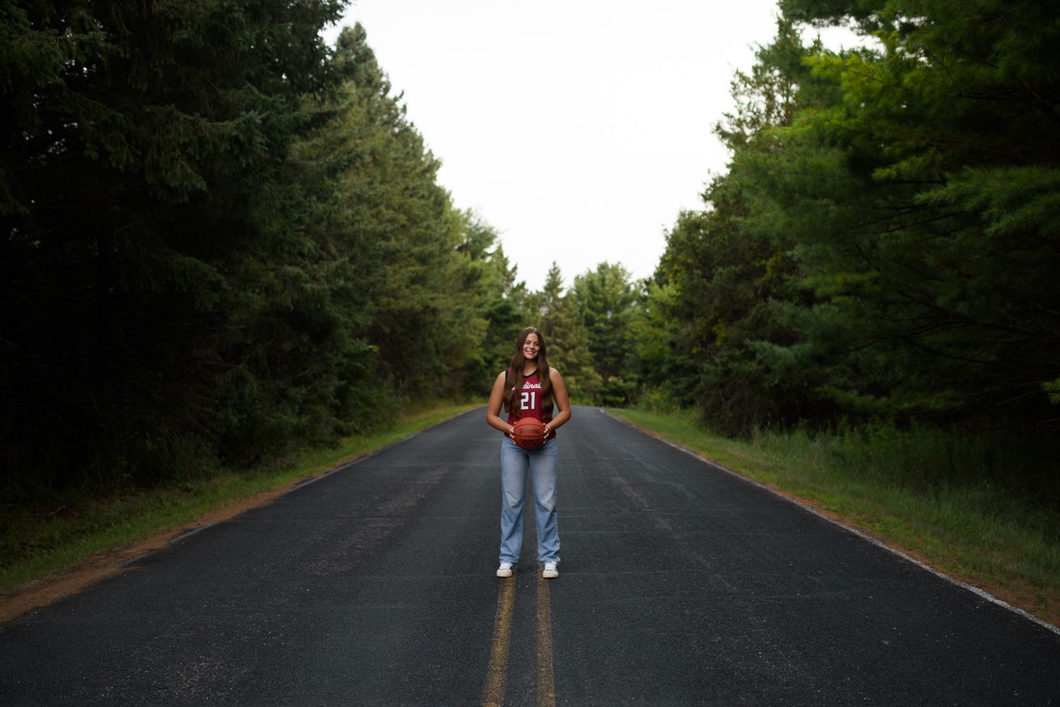 Young woman standing alone on a straight tree-lined road, centered between yellow dashes, wearing jeans and a dark top.
