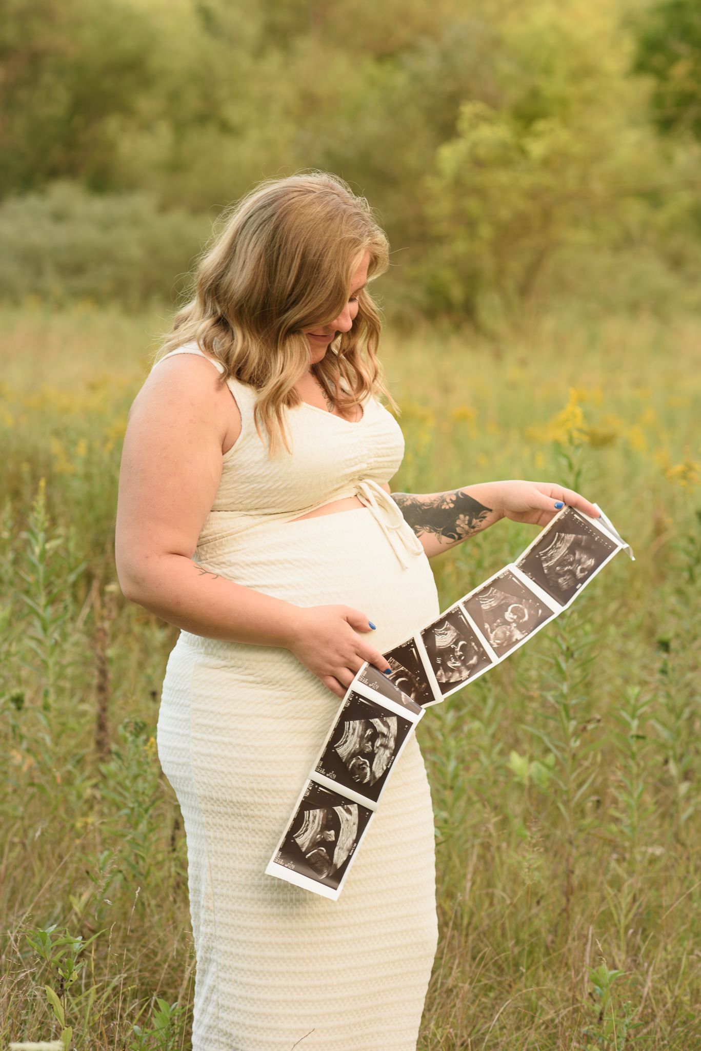 Pregnant woman in cream dress holding ultrasound photos in a sunlit field during a maternity photography session.