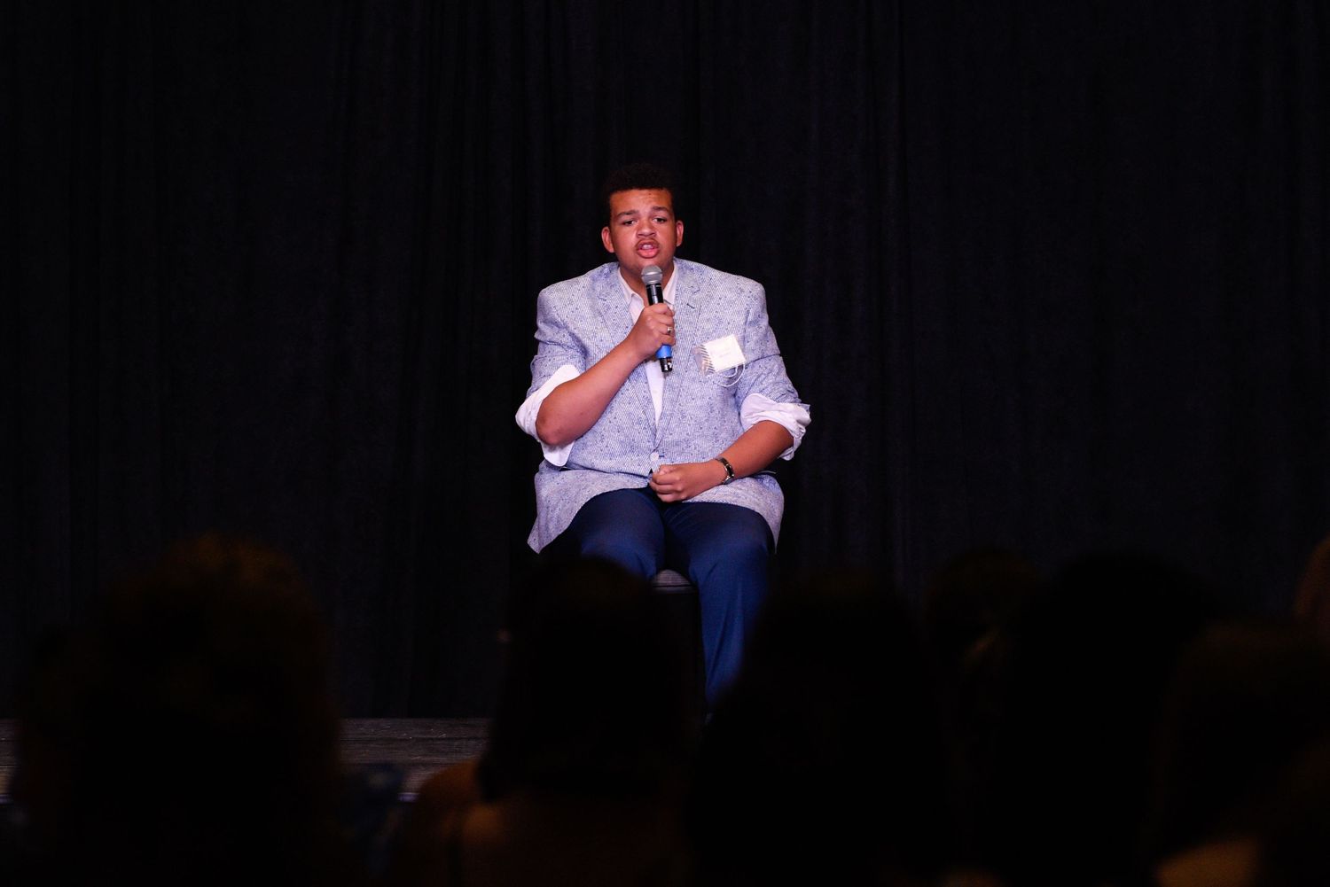 Man holding microphone speaking to audience while seated on stage at indoor event with dark backdrop.
