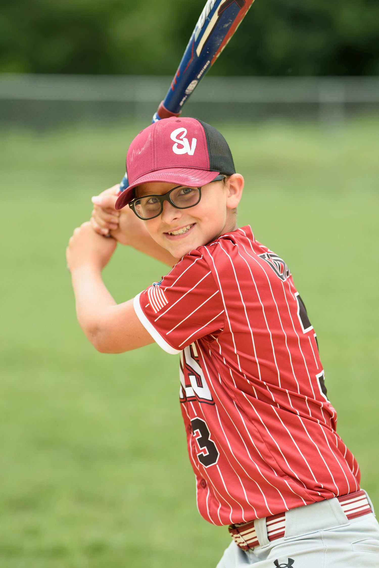 Smiling young baseball player wearing glasses in Cardinals uniform posed in batting stance outdoors.