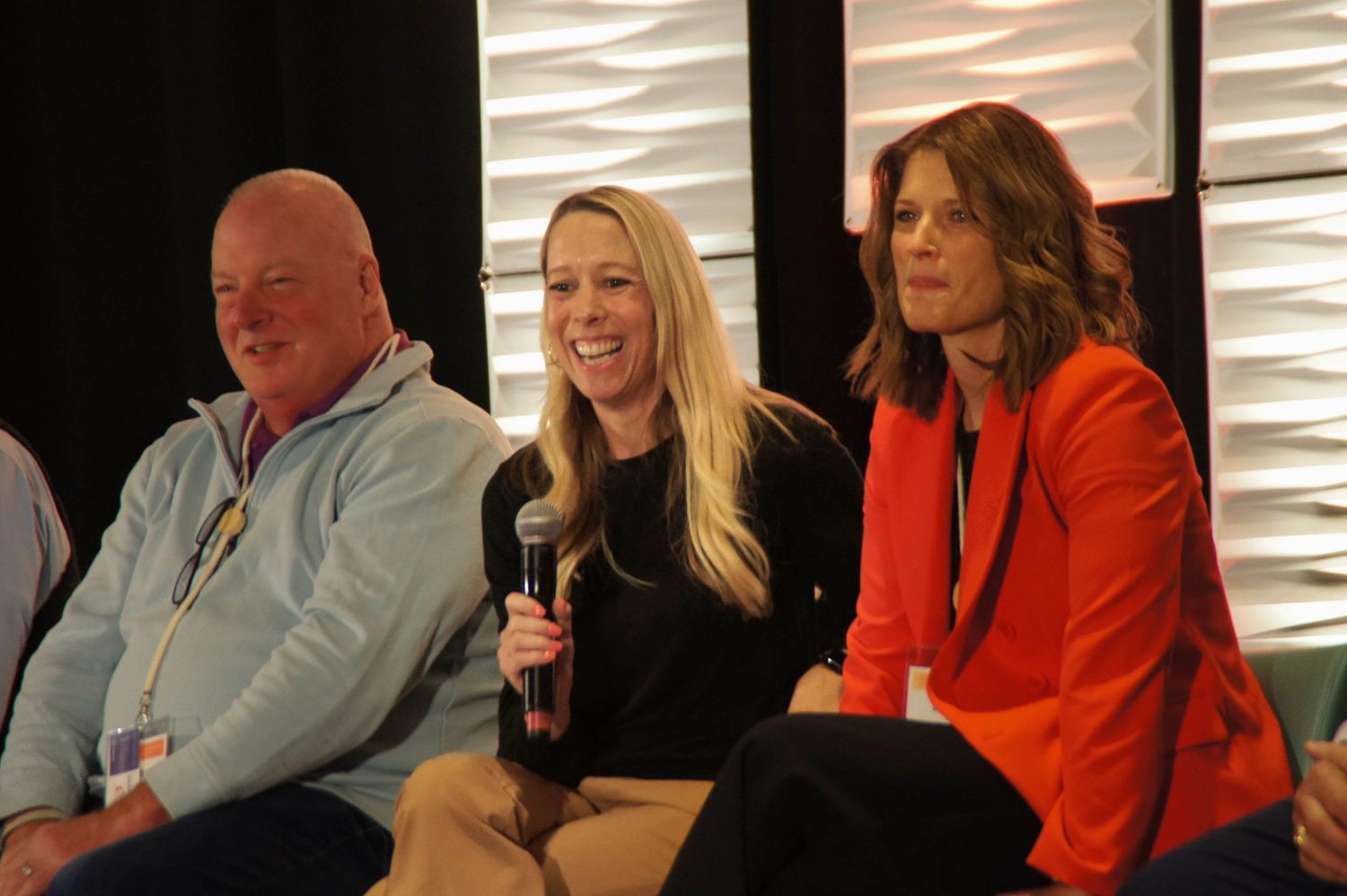 Three panelists smile during a conference discussion, with a woman holding a microphone in the center.
