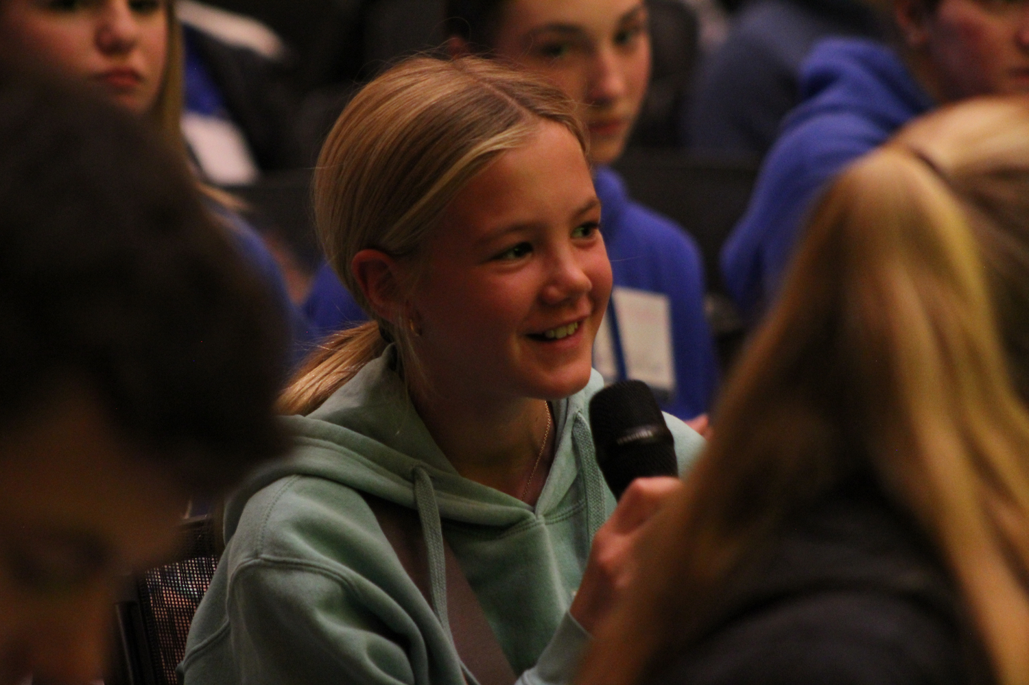 A smiling blonde girl holds a microphone during an indoor student panel or Q&A session.