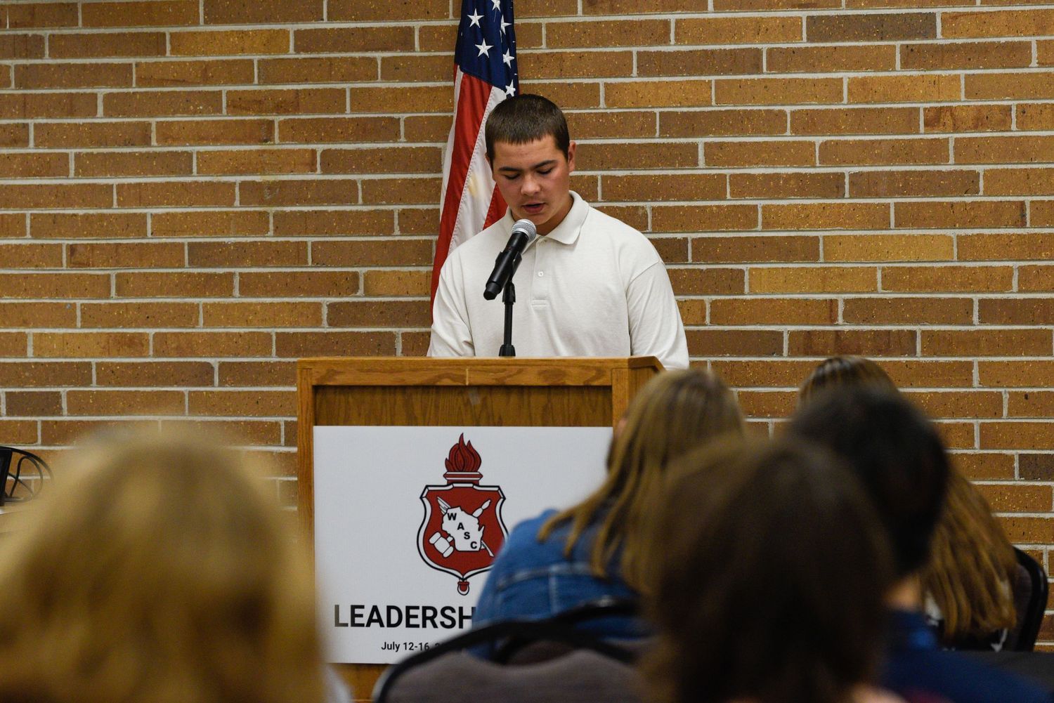 Student speaks at a leadership podium in front of a brick wall with an American flag during a school event.