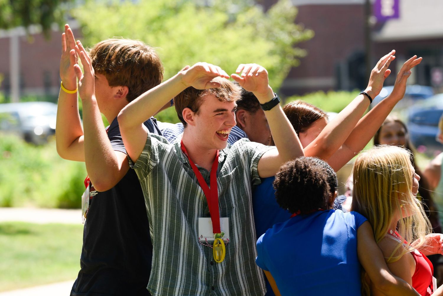 Students celebrate outdoors on a sunny day, raising hands and smiling, wearing lanyards at a campus event.