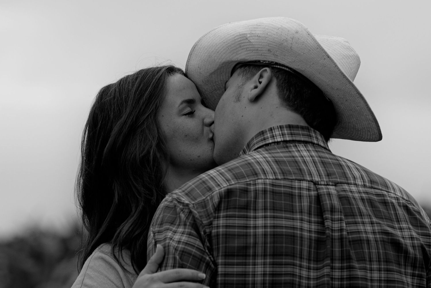 Black and white photo of a couple sharing a romantic kiss, man wearing a cowboy hat and plaid shirt outdoors.