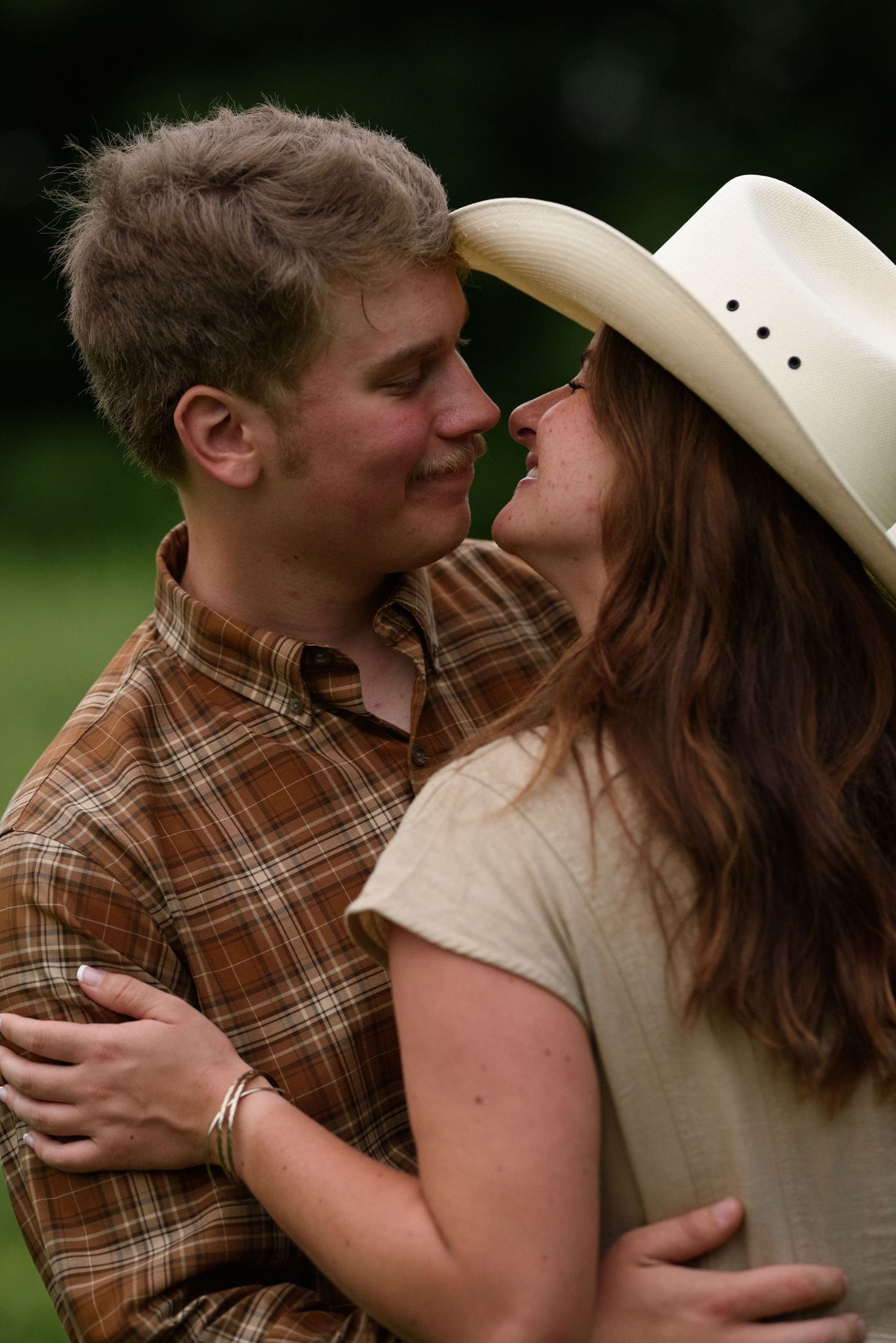 A young couple in plaid and cowboy hat embrace and smile at each other outdoors.