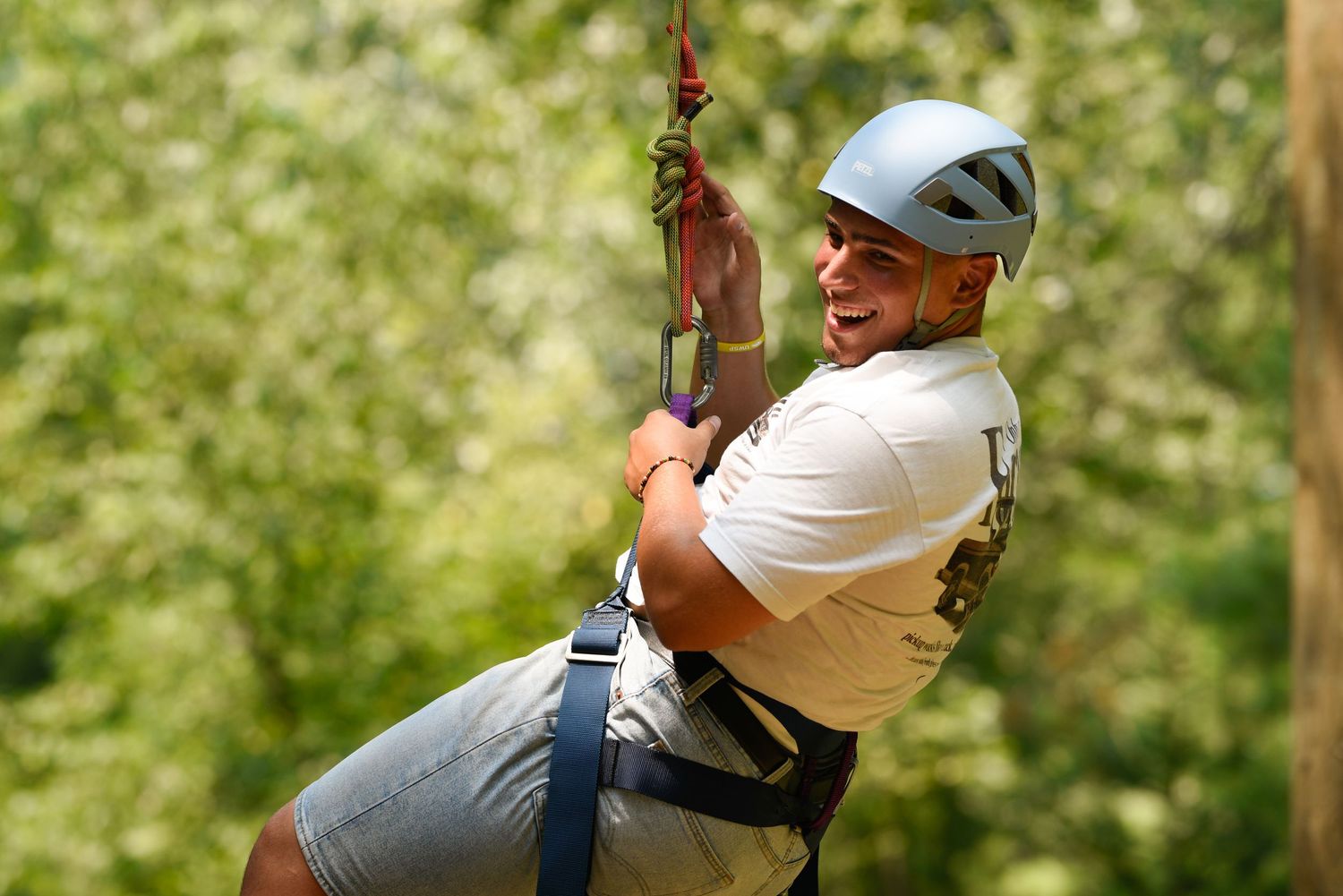 Person wearing helmet and harness smiling while zip-lining through a lush green forest.