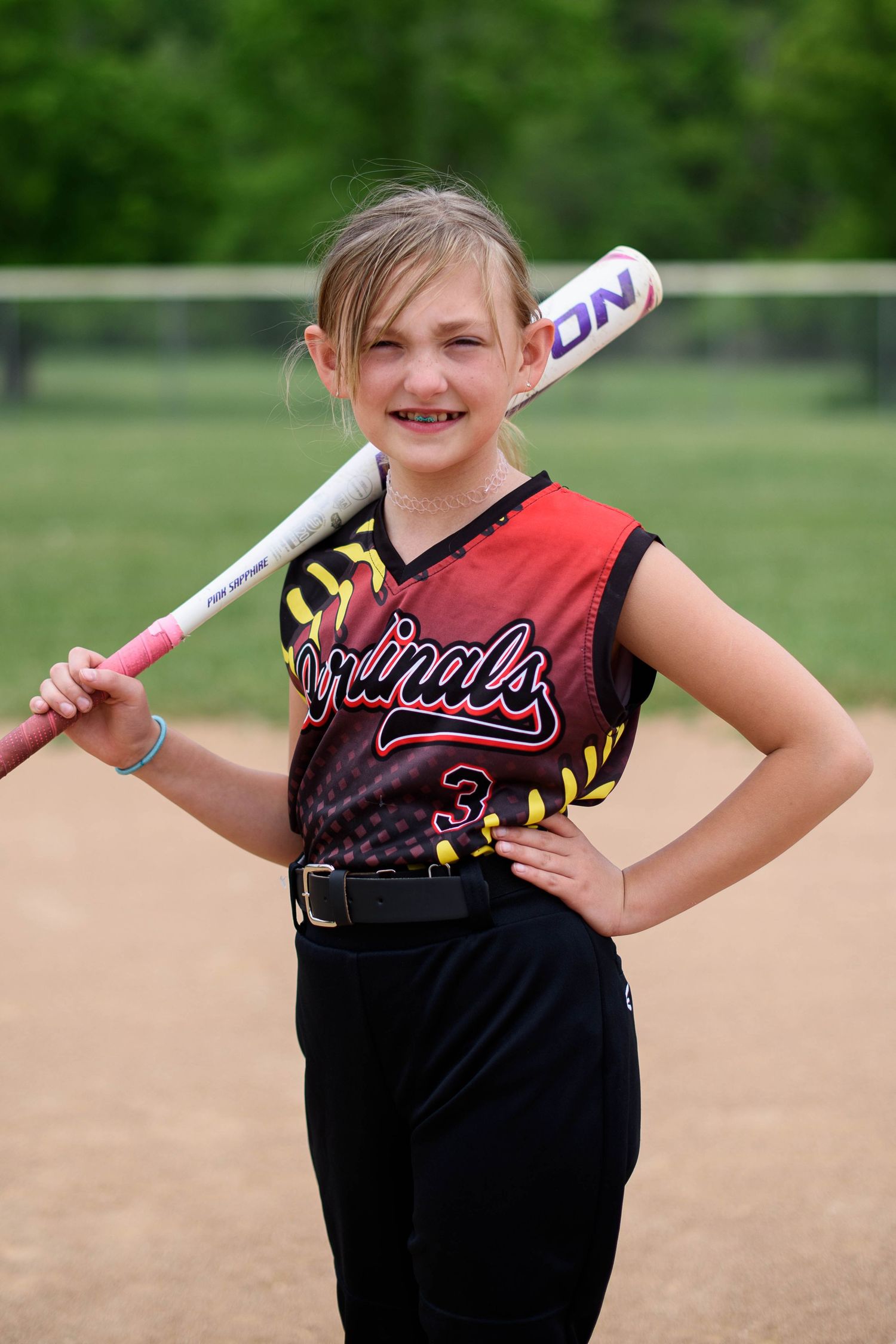 Young girl in Cardinals softball uniform holding a bat over her shoulder, smiling on a baseball field.
