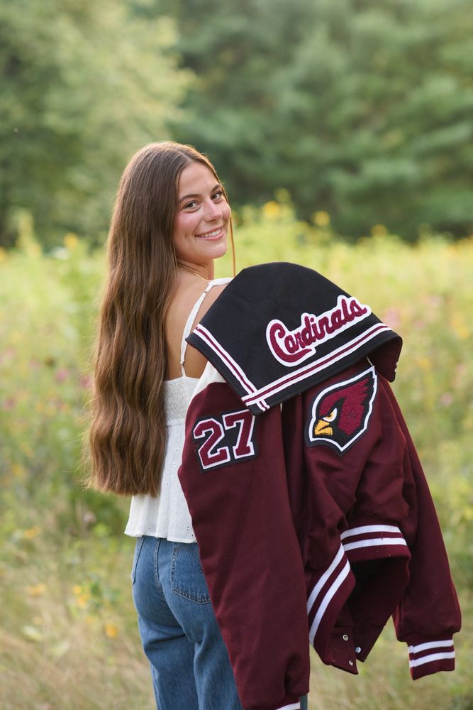 Smiling teen girl holding a Cardinals varsity jacket numbered 27 outdoors in a wildflower field.