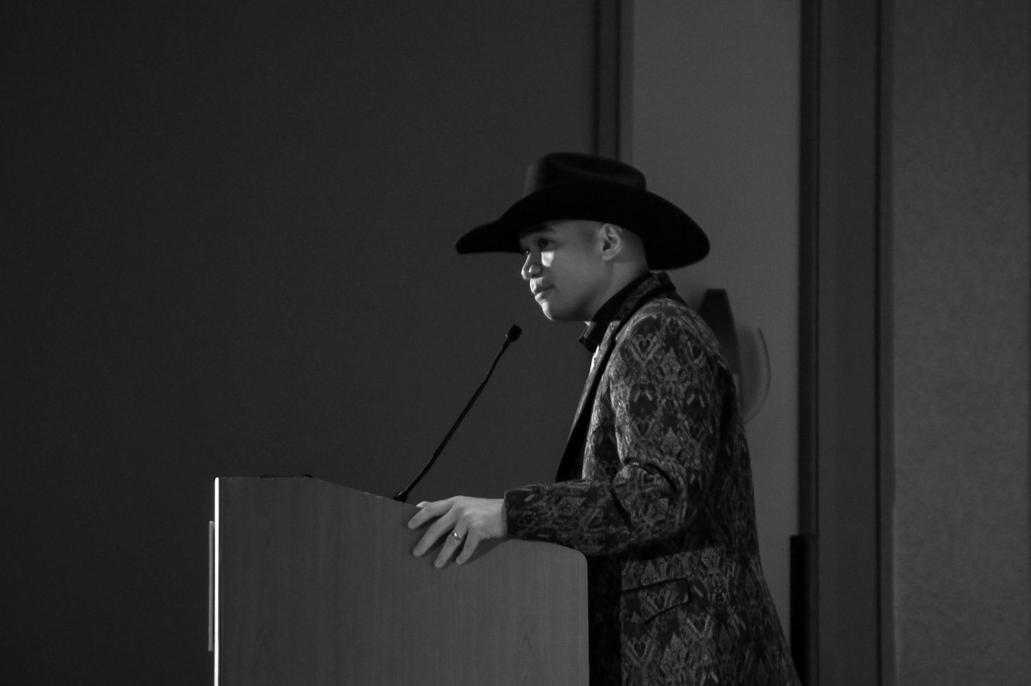 Black and white photo of a man wearing a cowboy hat speaking at a podium with microphone.