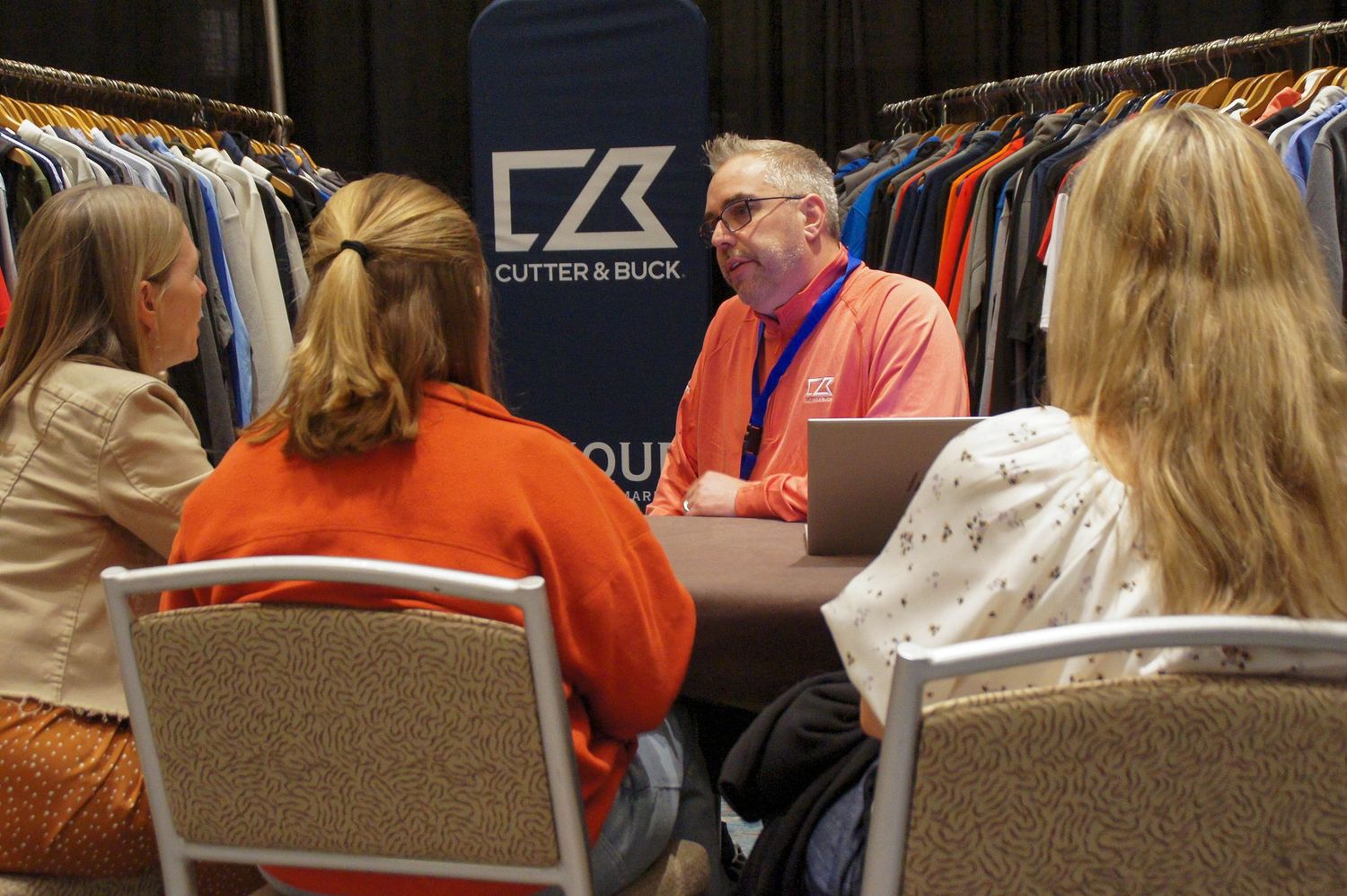 Man presenting Cutter and Buck apparel line to two women at a trade show booth.
