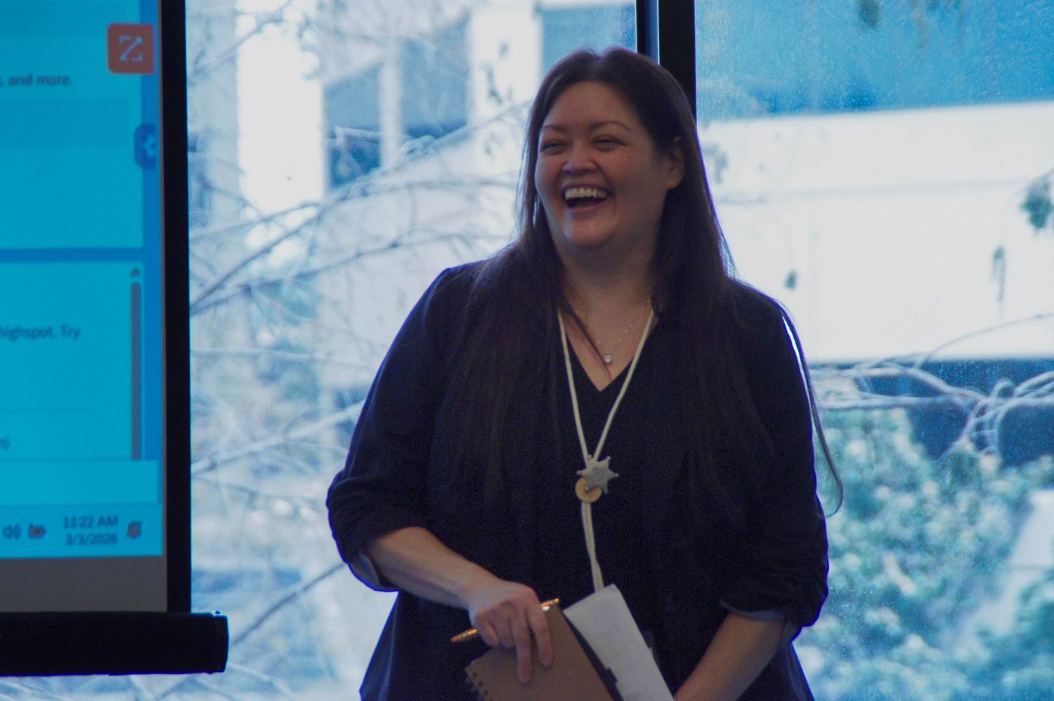 Smiling woman in navy blouse holding papers, standing near a presentation screen in a modern office.