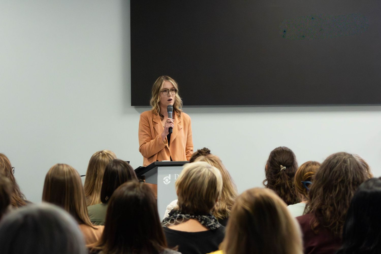 Woman in orange blazer speaks at podium with microphone to seated audience in modern conference room.