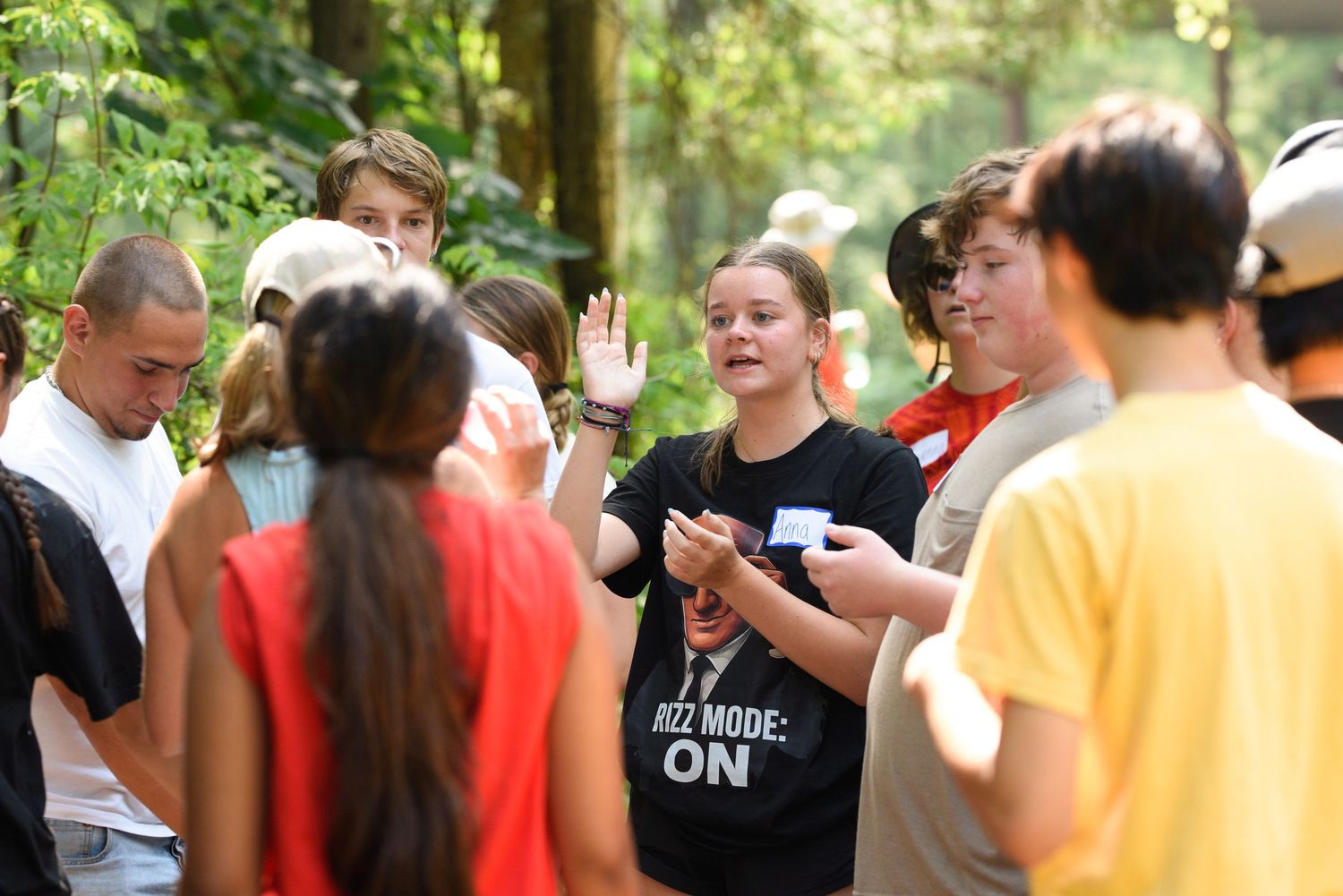 Students gather outdoors in a forest setting, listening to a peer in a black shirt during a group activity.