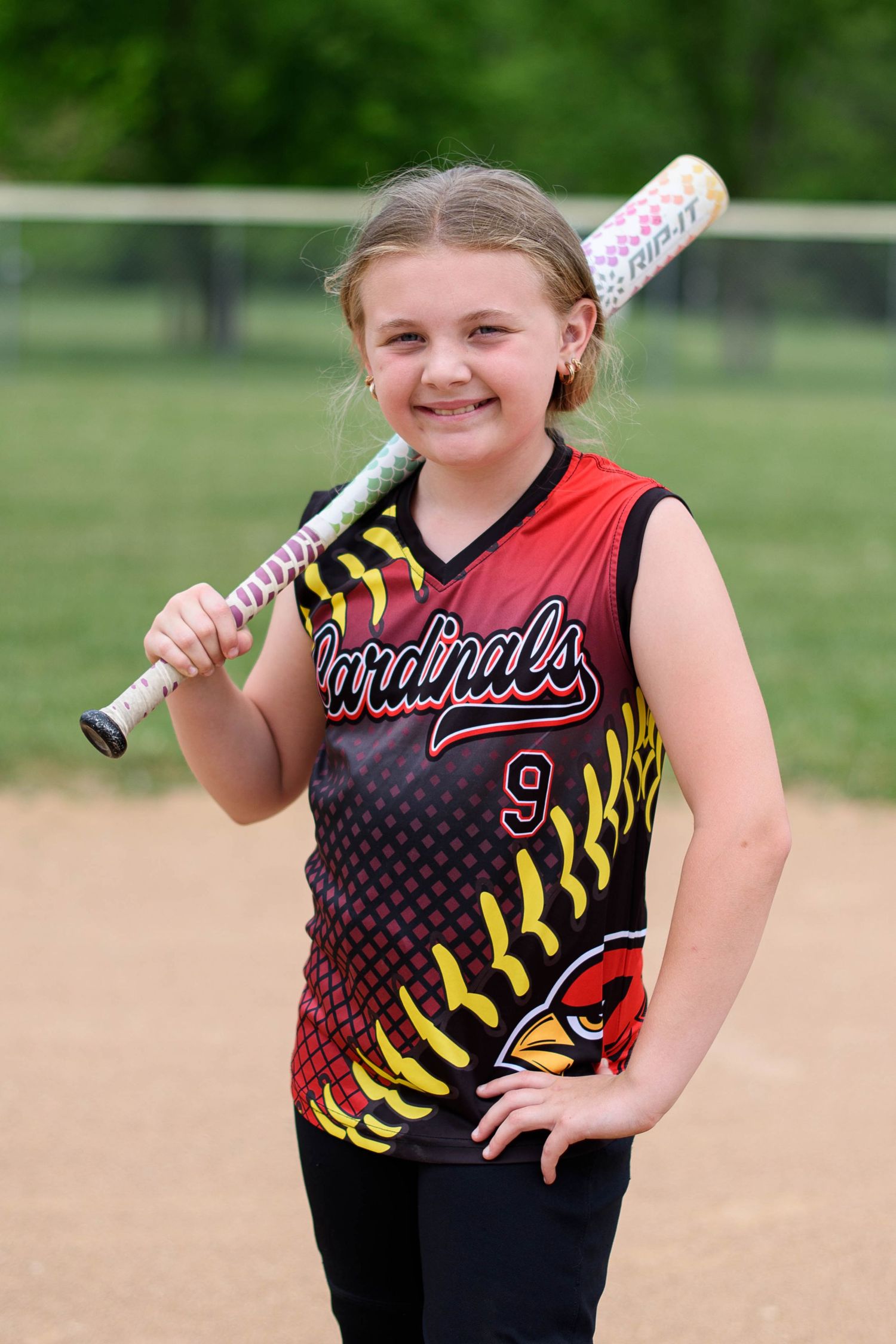 Young Cardinals softball player #9 smiling and holding a colorful bat on her shoulder on a dirt field.