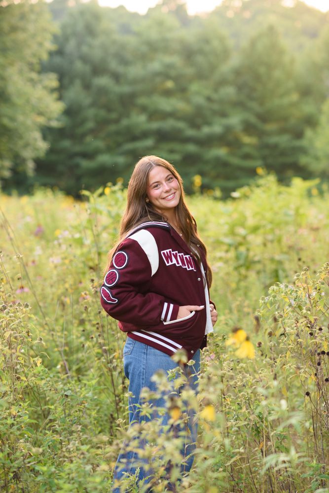Smiling teen girl in maroon varsity jacket standing in wildflower field with green trees in background.