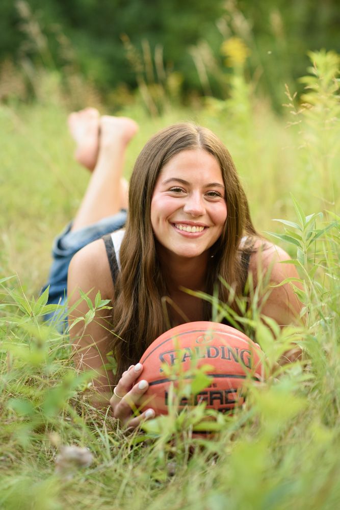Smiling teenage girl lying in green grass holding a Spalding basketball during an outdoor senior portrait session.