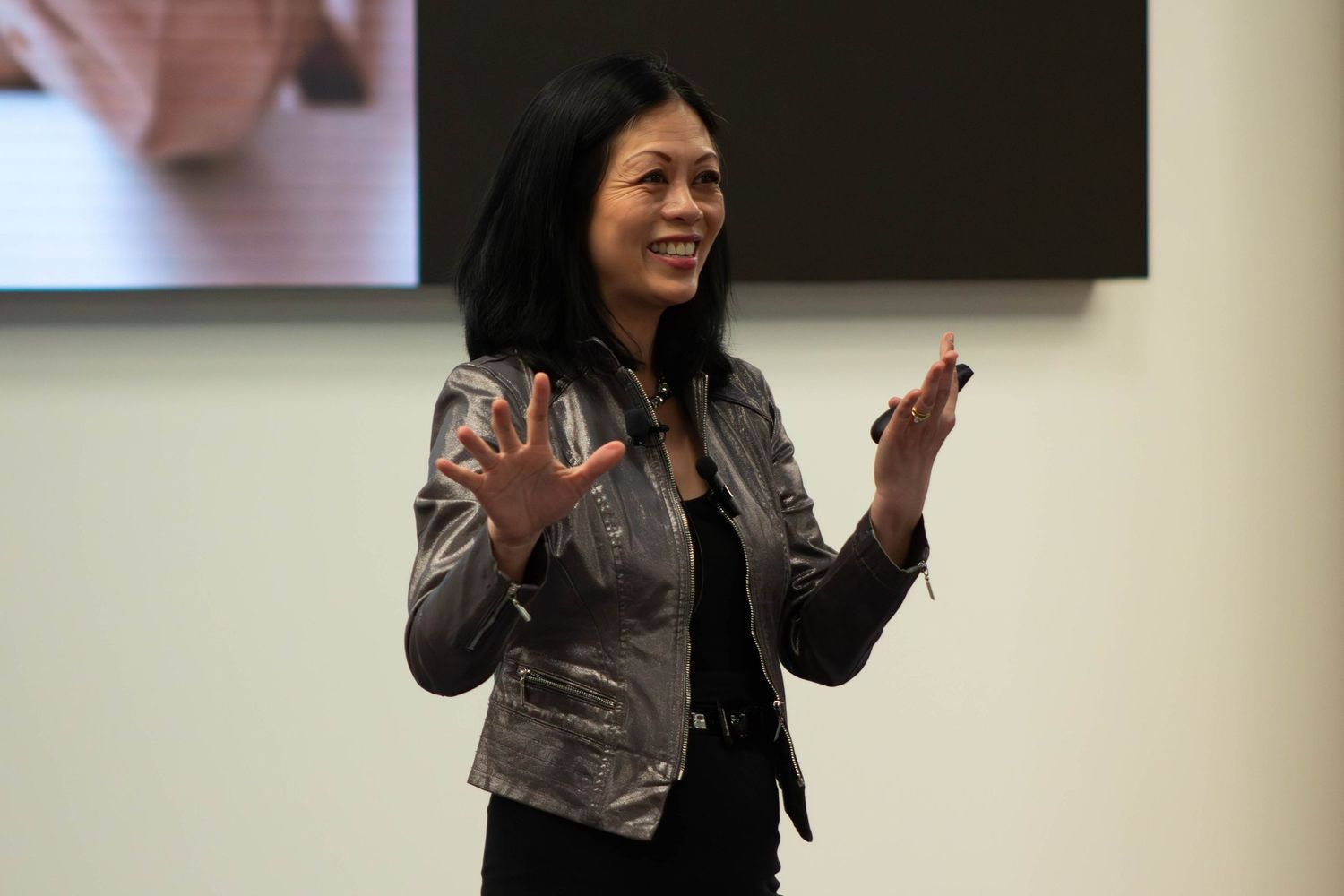 Woman in silver jacket gesturing while speaking at a professional presentation event.