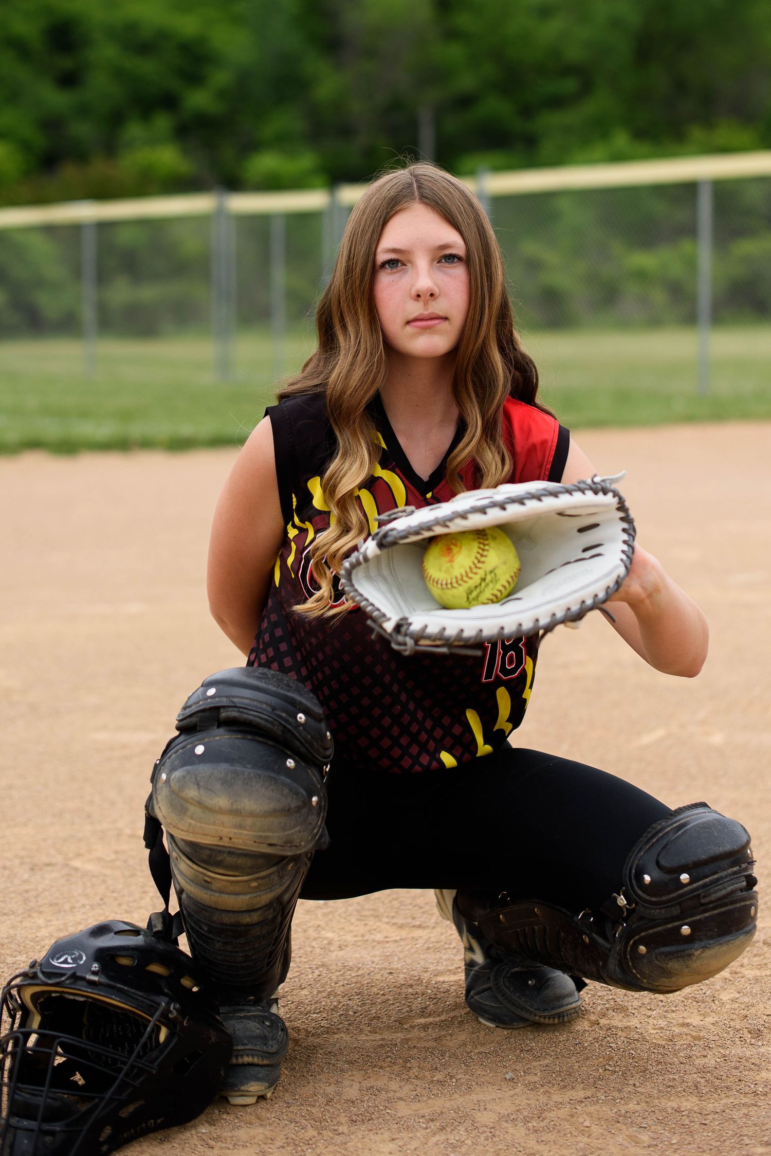 Young female softball catcher in uniform kneeling on dirt field, holding yellow softball in white catcher's mitt.