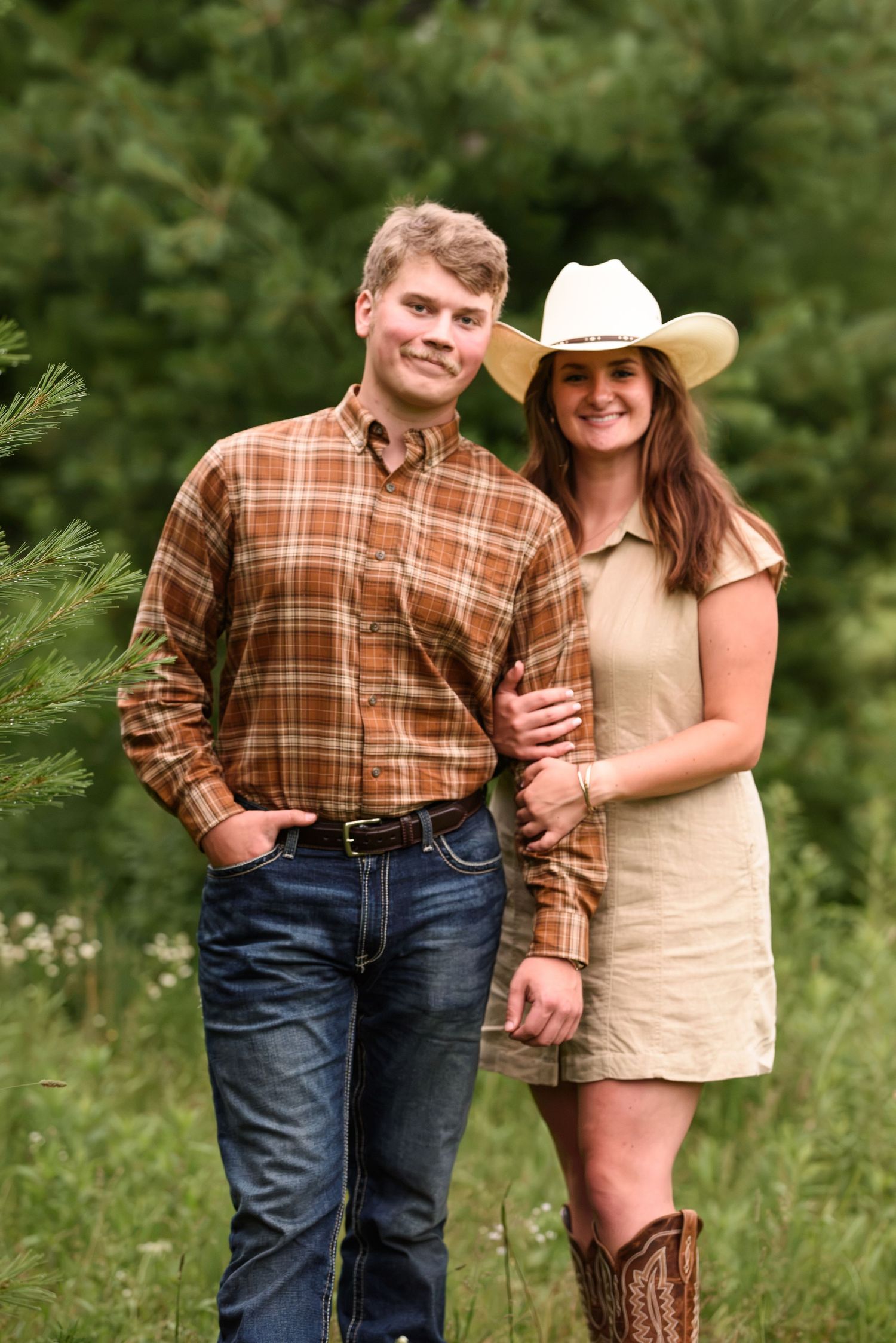 Couple standing by pine tree, woman in cowboy hat and boots, man in plaid shirt and jeans, smiling outdoors.