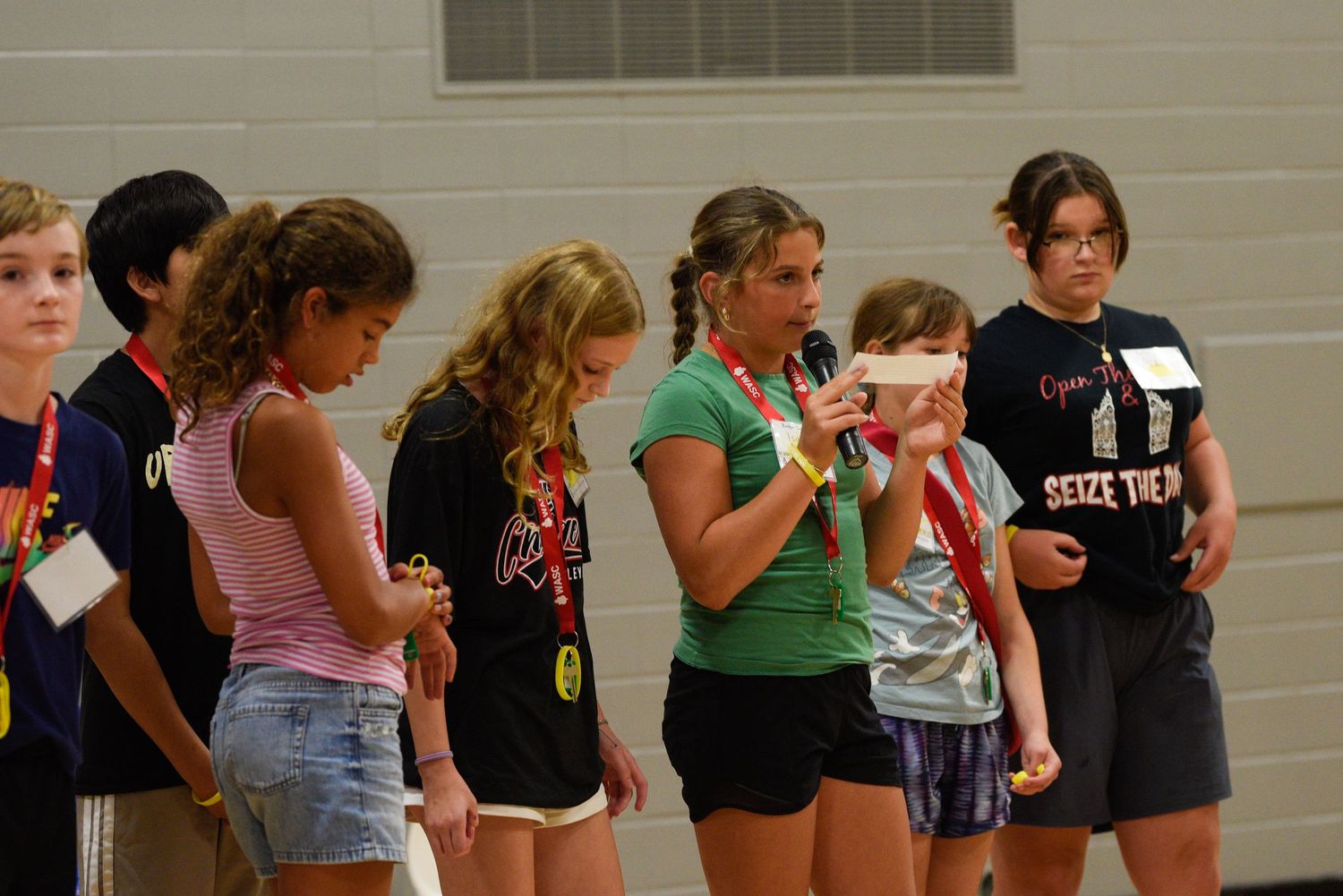 Group of children standing in a line indoors at a school event, with a girl speaking into a microphone.