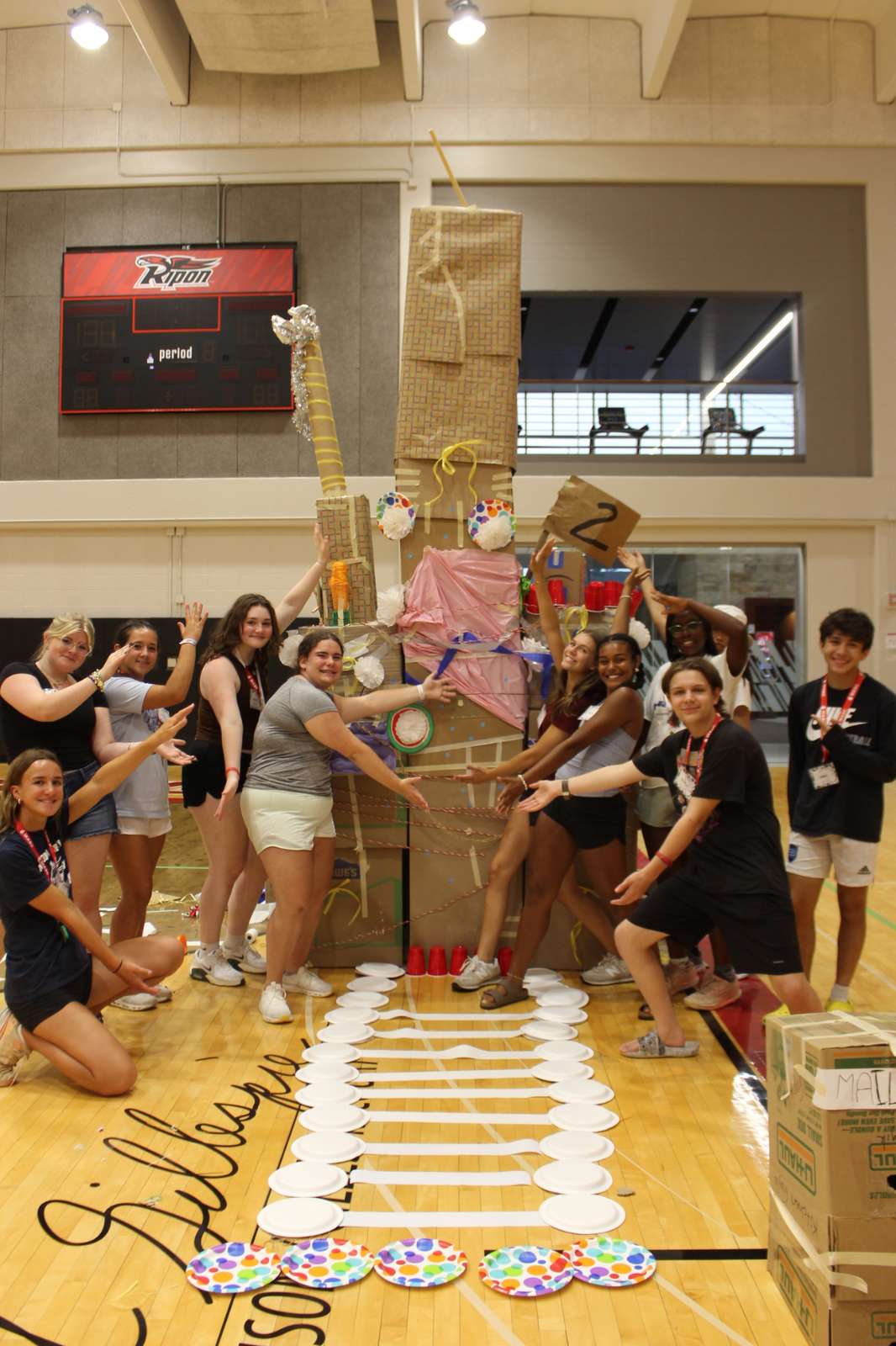 Students pose proudly beside a tall cardboard sculpture built in a gymnasium during a creative team challenge.