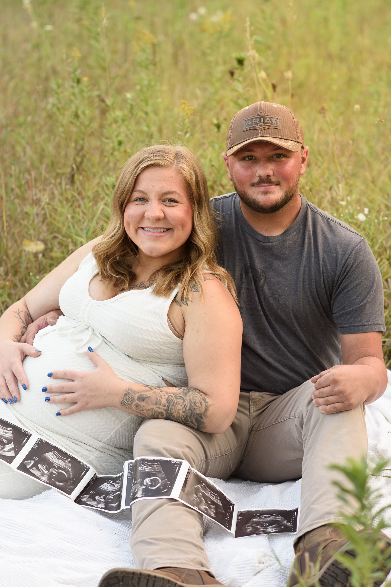 Smiling pregnant couple sitting in a field with ultrasound photos, celebrating their pregnancy during an outdoor maternity photoshoot.