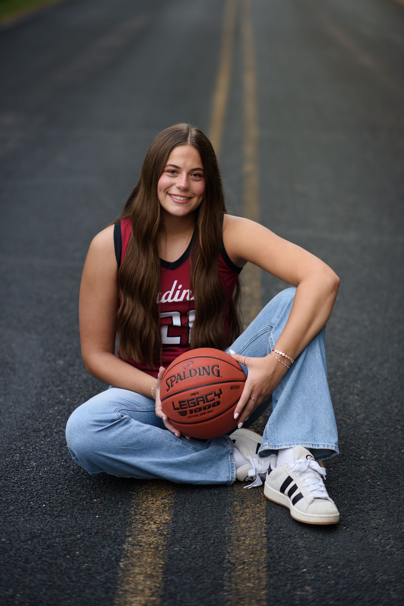 Young female basketball player sitting cross-legged on a road holding a Spalding basketball, wearing a Cardinals jersey.