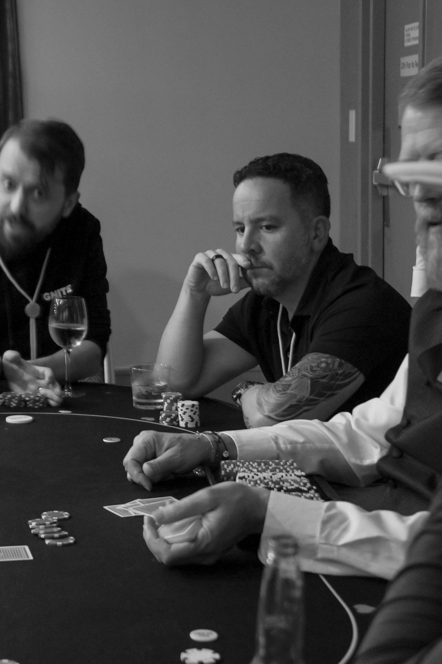 Tattooed man contemplating his hand at a black and white poker table.