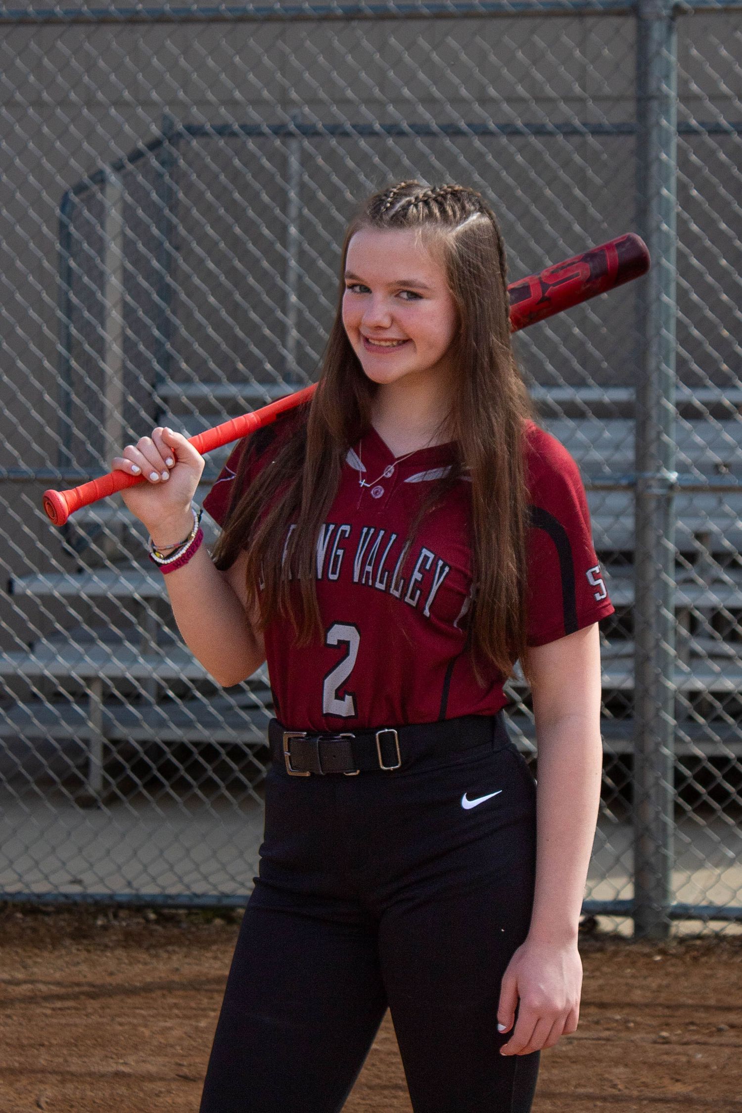 Spring Valley softball player #2 poses with a red bat over her shoulder in a maroon jersey on the field.