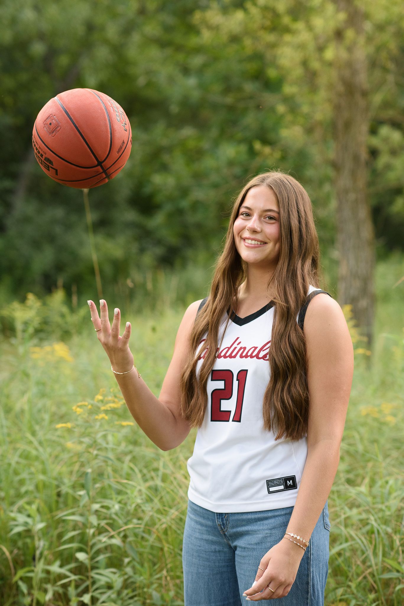 Smiling teen girl in Cardinals basketball jersey #21 tossing a basketball outdoors.