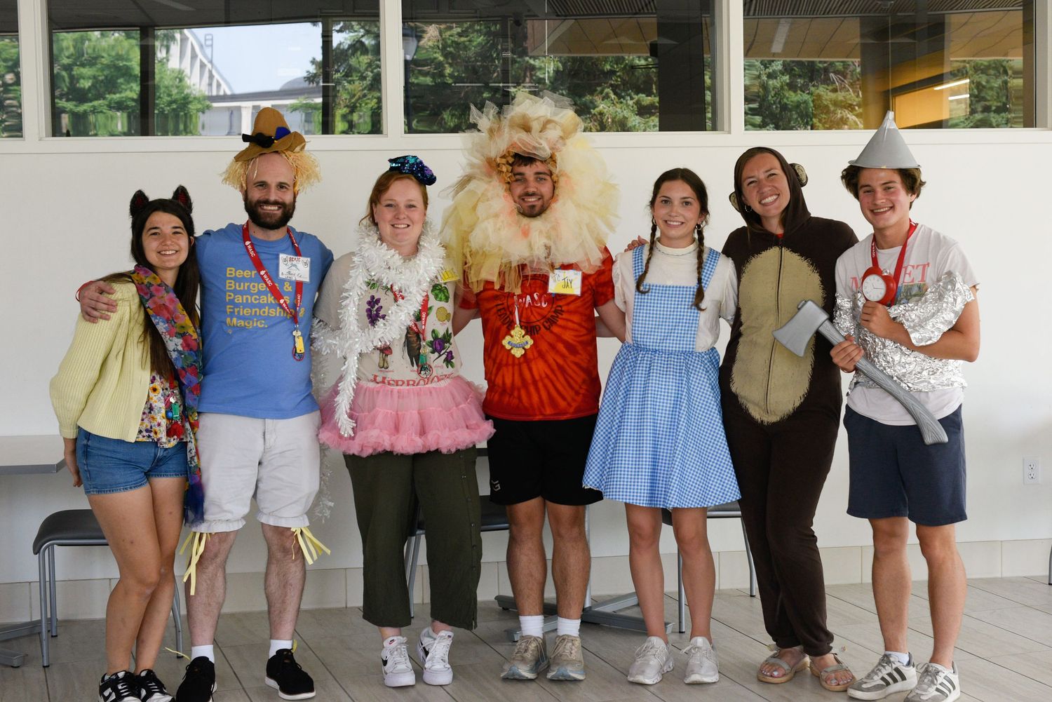 Group of seven people in Wizard of Oz costumes posing together indoors, including Dorothy, Lion, and Scarecrow.