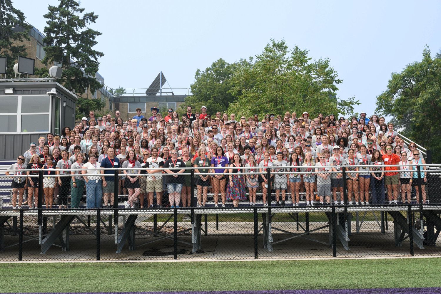 Large group of students posing on bleachers outdoors on a sunny day at a school athletic field.