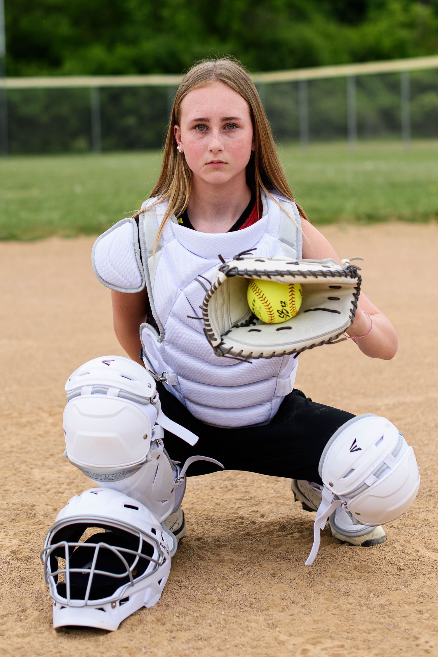Young female softball catcher in white gear crouching on dirt field holding yellow ball in glove.