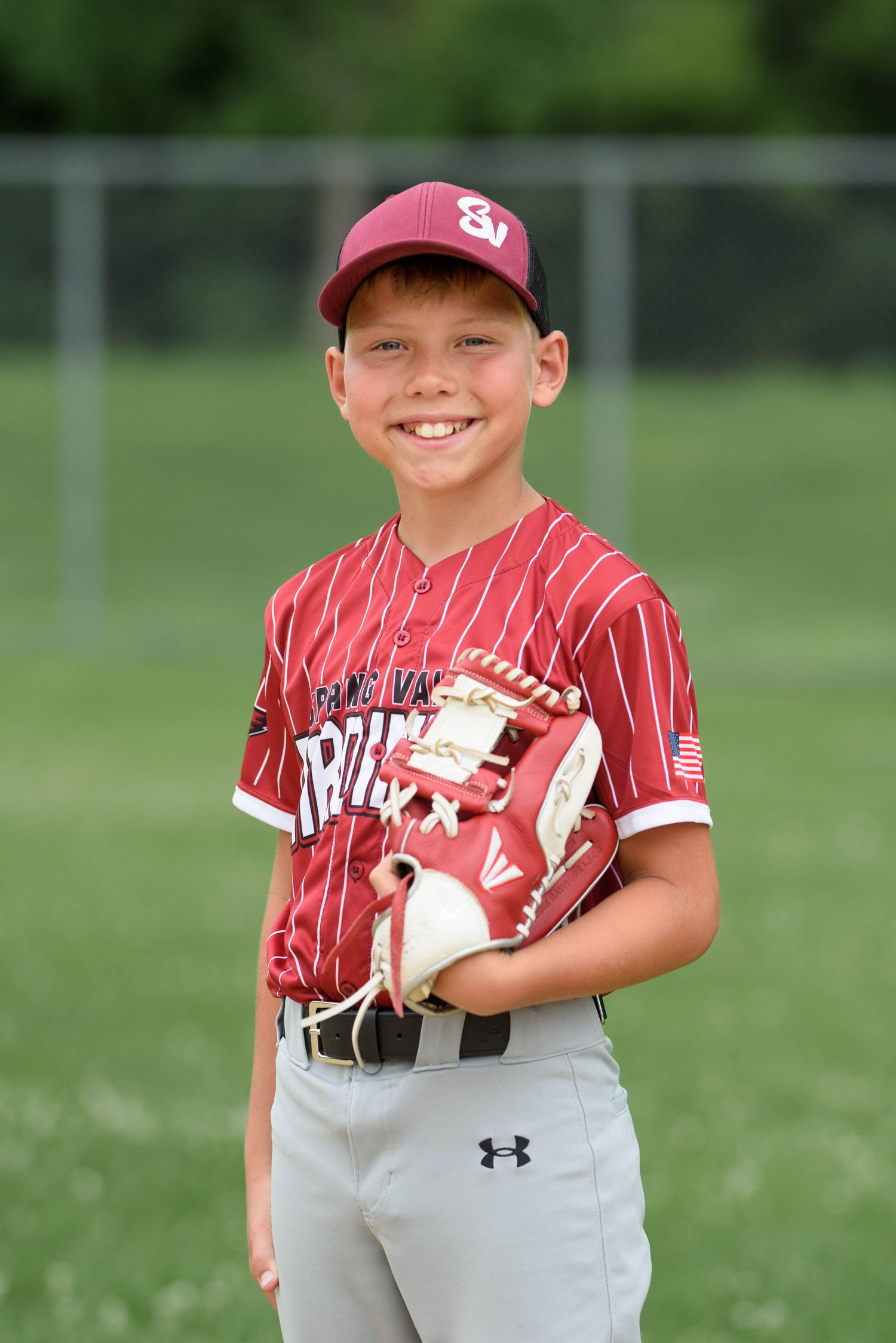 Young baseball player in maroon pinstripe uniform smiling on field, holding glove, wearing matching cap.