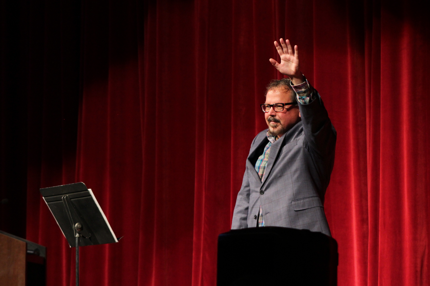 Male speaker raises hand while presenting on stage before a red curtain at a conference.
