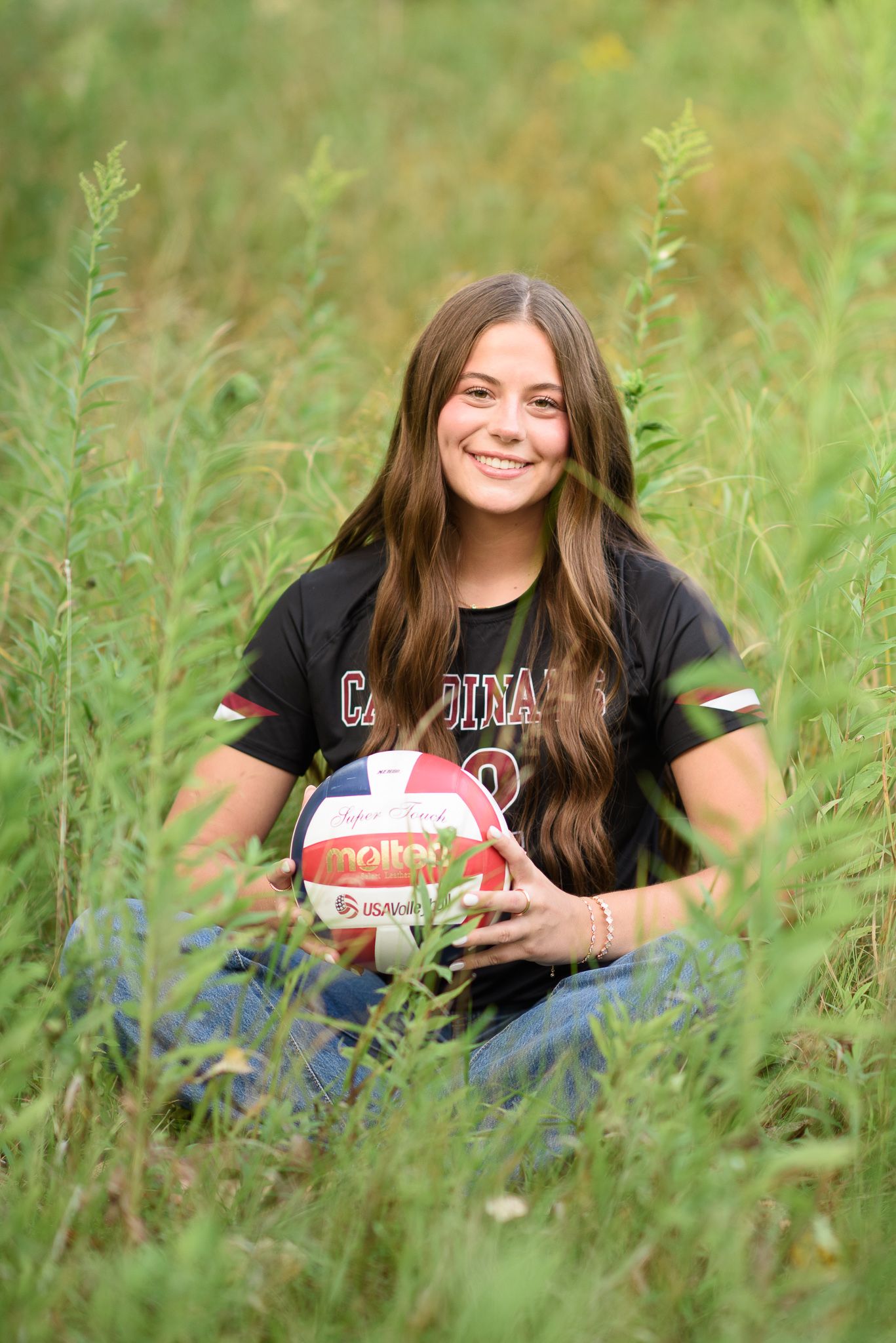 Teen girl in Cardinals volleyball jersey sitting in tall grass holding a Molten volleyball.