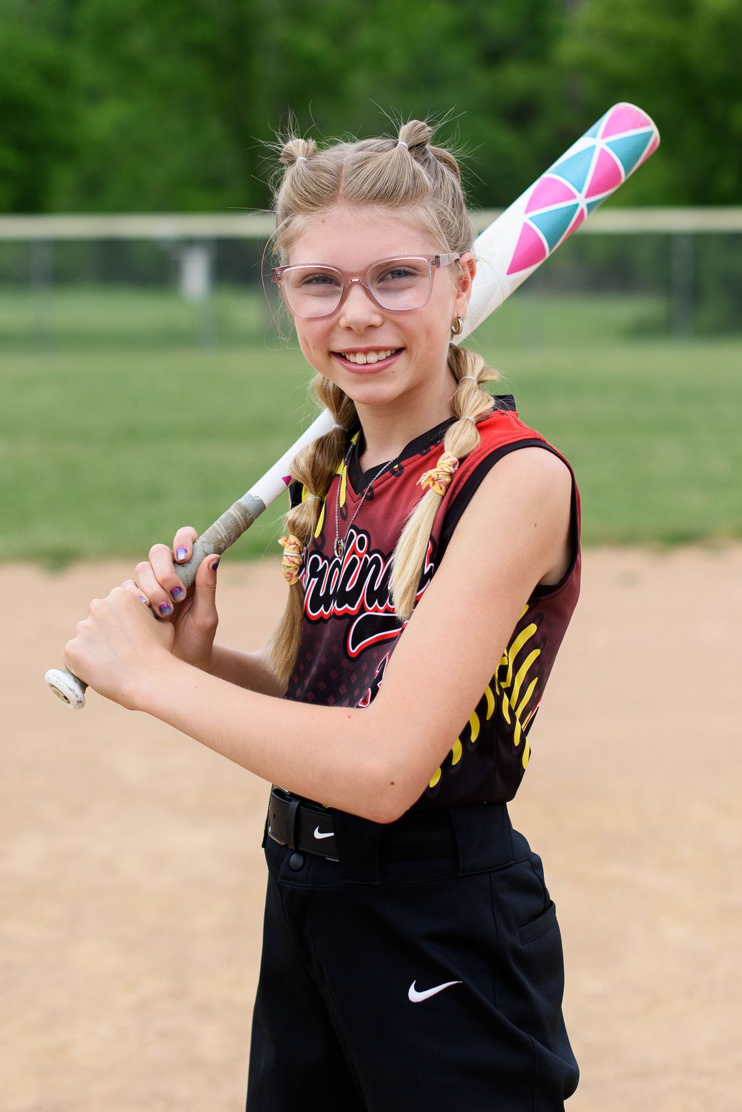 Young Cardinals softball player smiling in glasses, holding a pink and white bat in a batting stance.