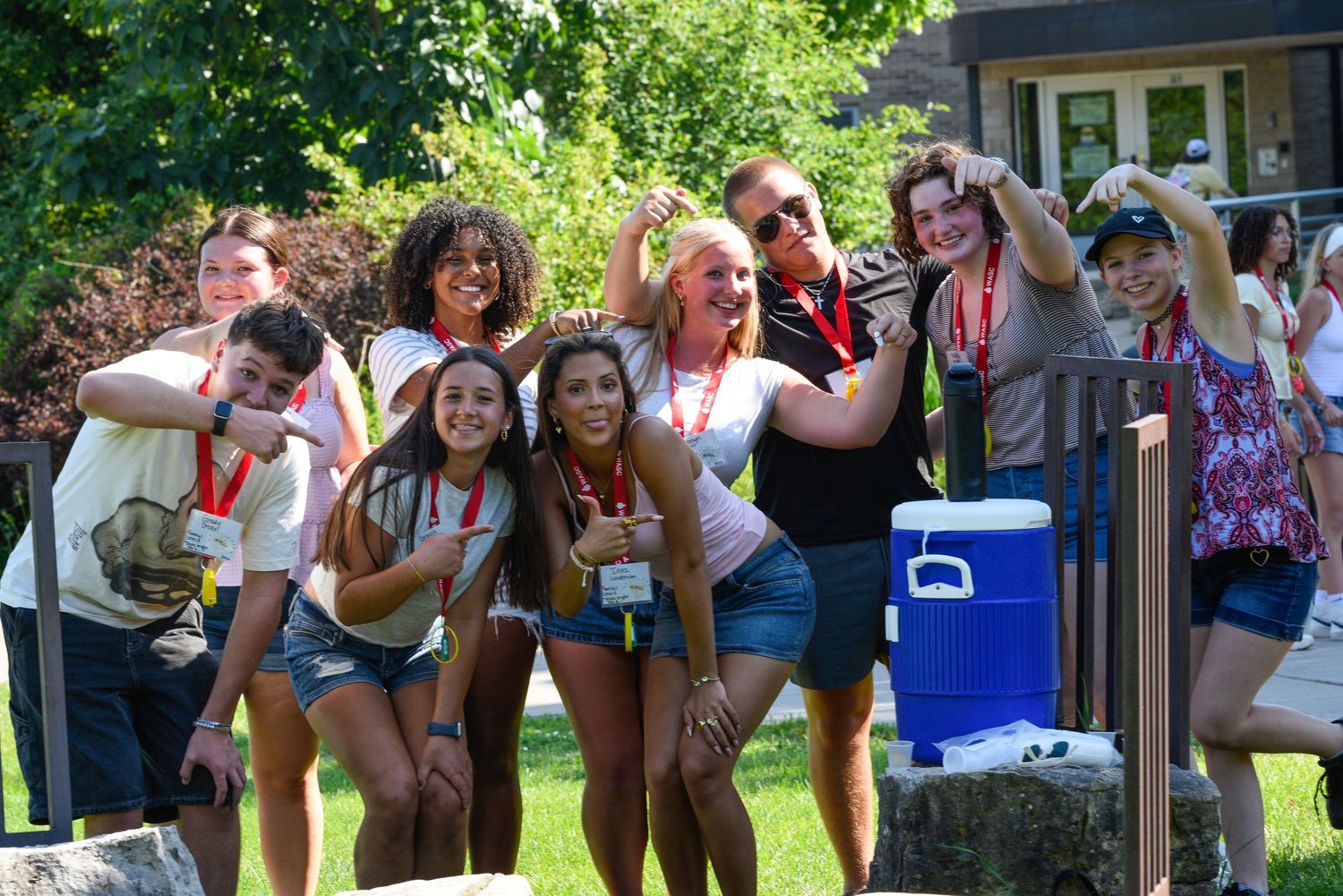 Group of smiling teens posing outdoors at summer camp on a sunny day with trees and a cooler nearby.