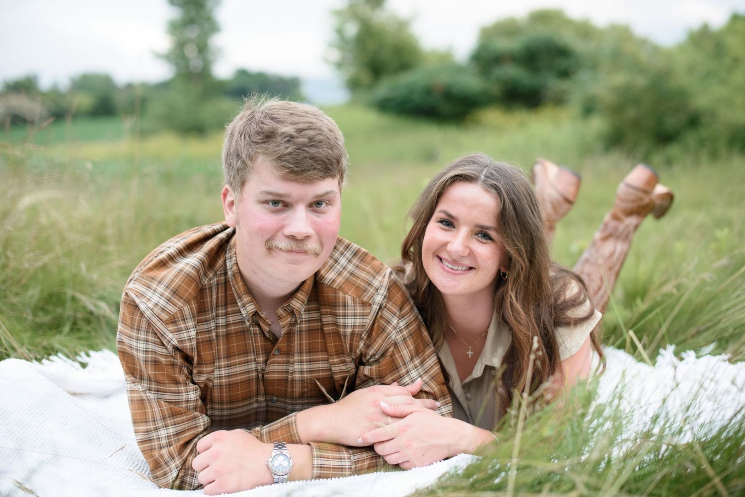 Happy young couple lying on a blanket in a grassy field, smiling at camera in matching brown plaid shirts.