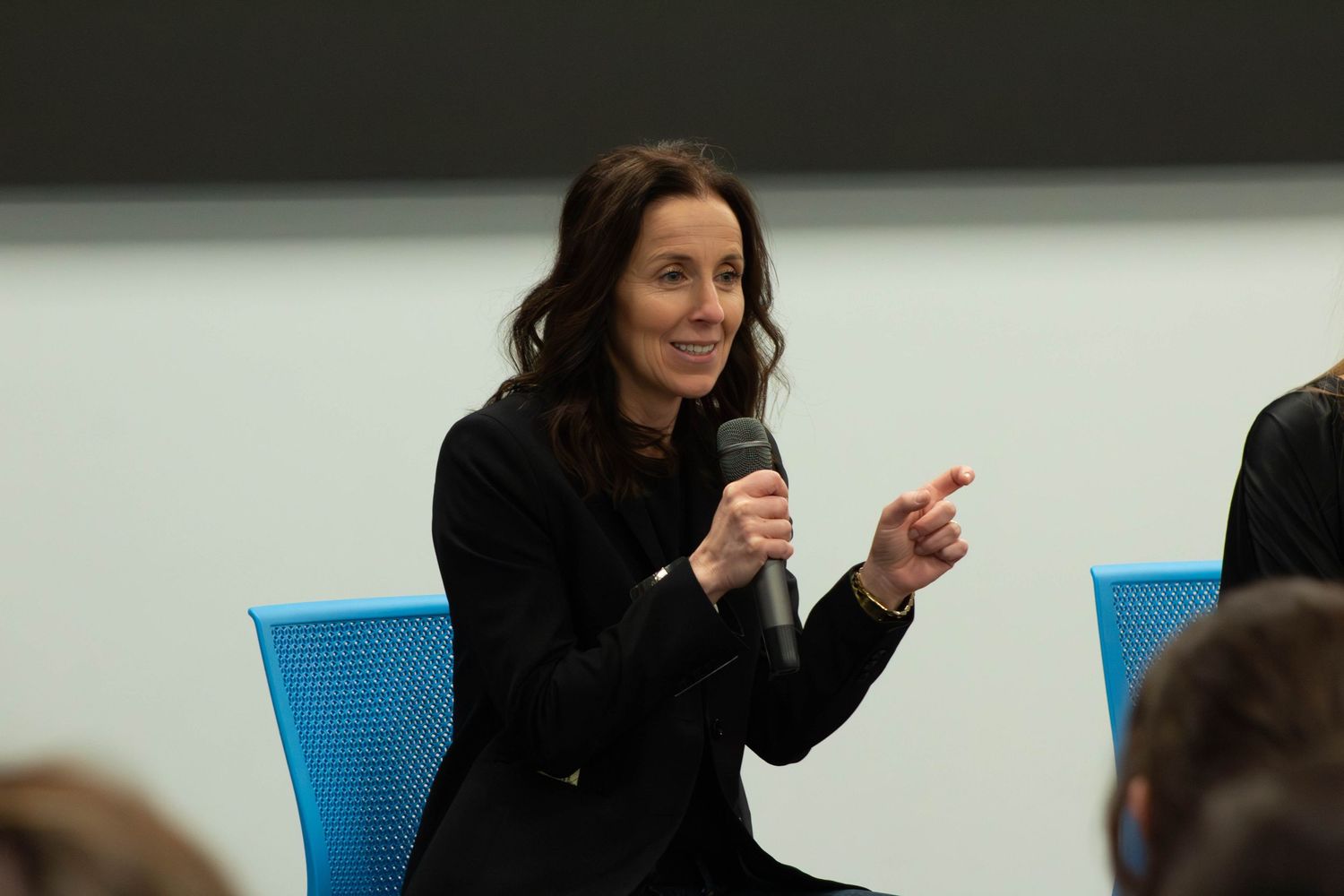 Woman in black blazer speaking into microphone, pointing finger during panel discussion event.