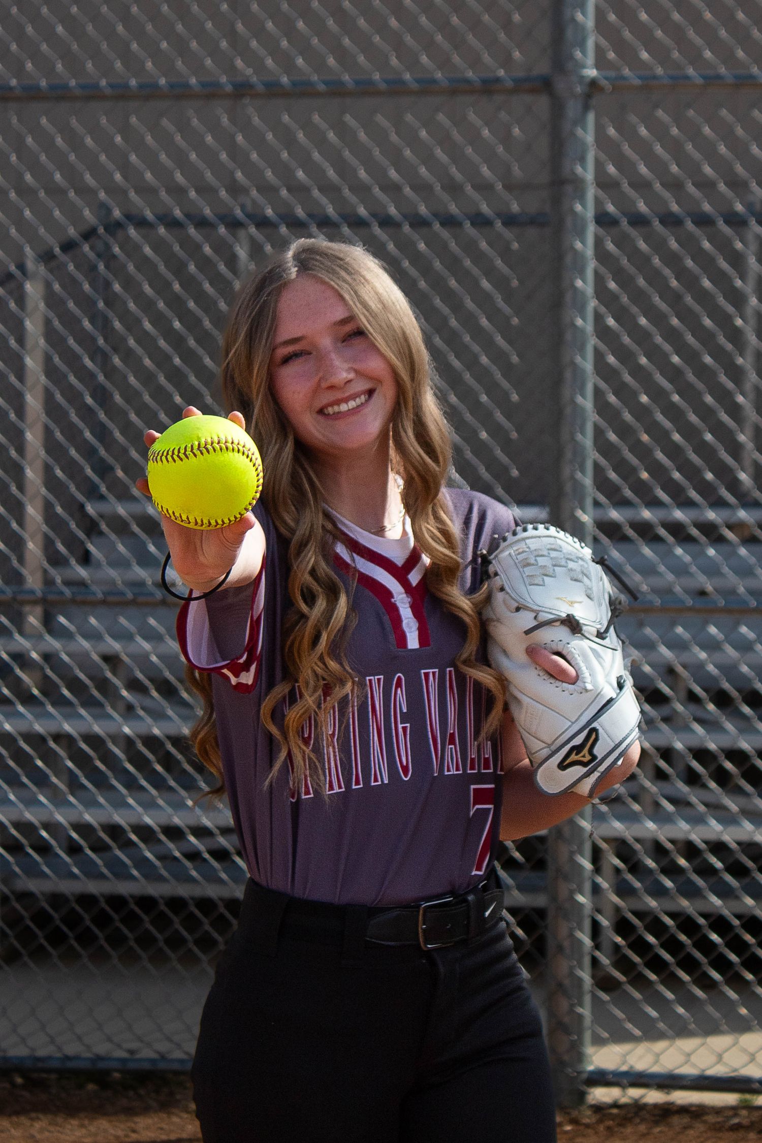 Spring Valley softball player #7 smiles holding a yellow softball and glove in a maroon jersey.