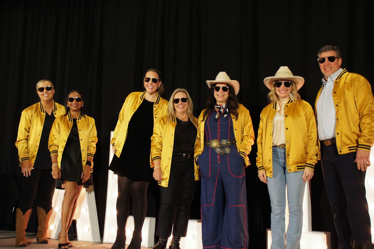 Group of seven people posing on stage wearing yellow varsity jackets and cowboy hats at ALL IN event.