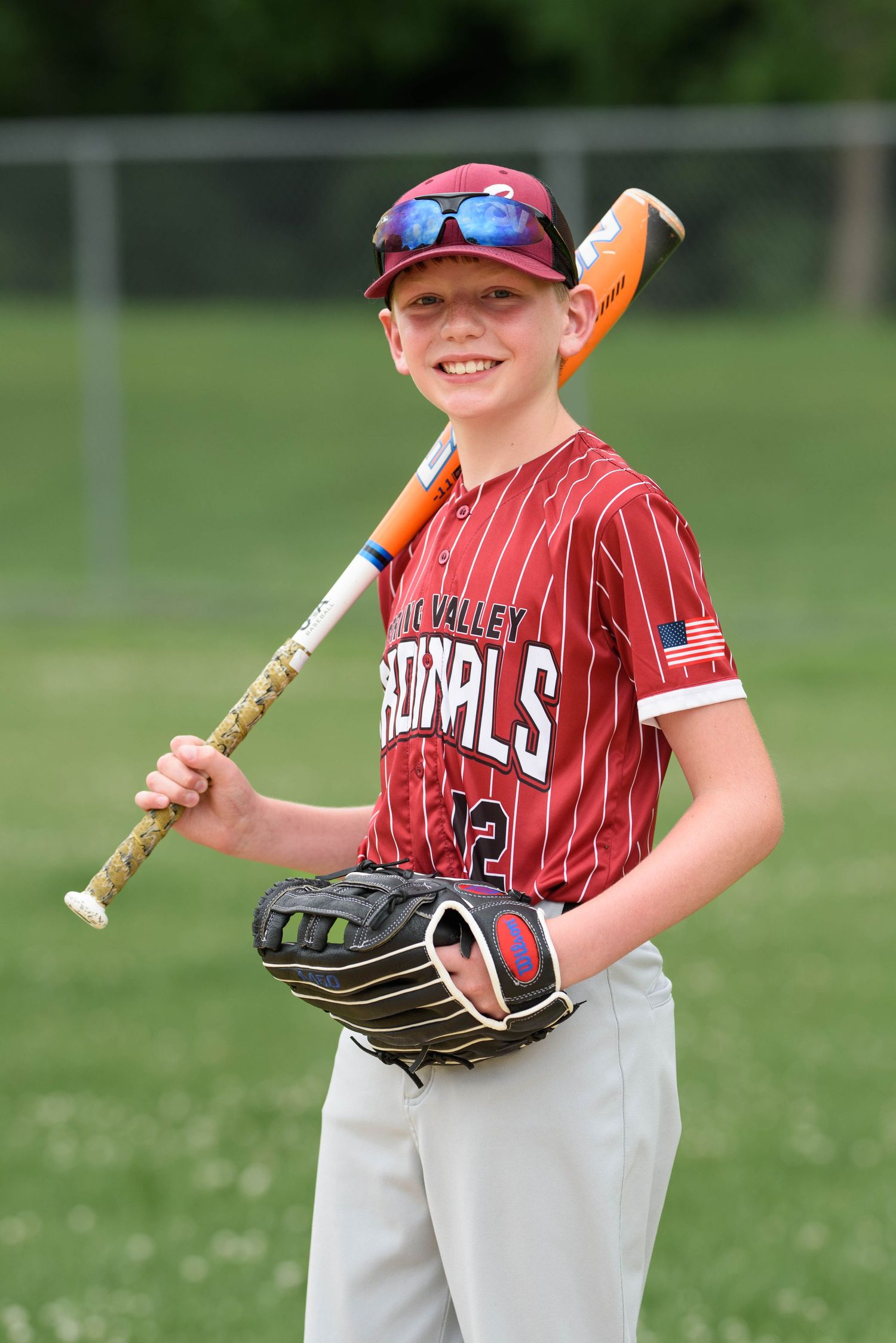 Smiling youth Cardinals baseball player #2 poses with bat and glove wearing sunglasses on green field.