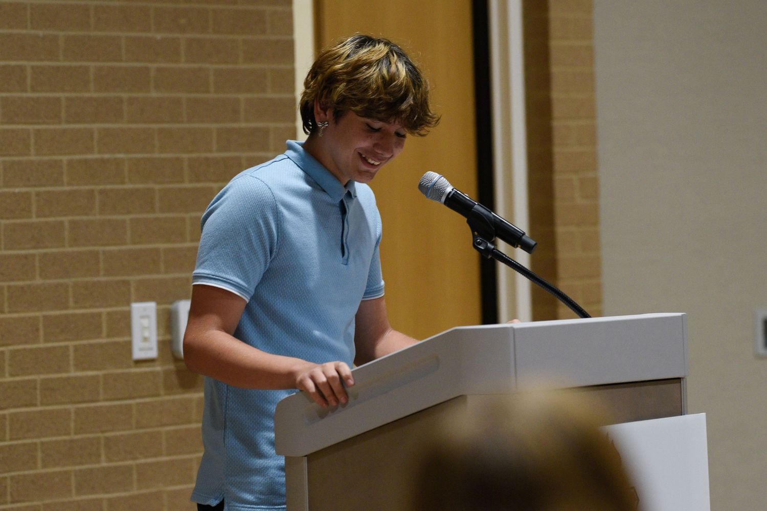Young student in blue polo shirt speaking at podium with microphone during school presentation event.
