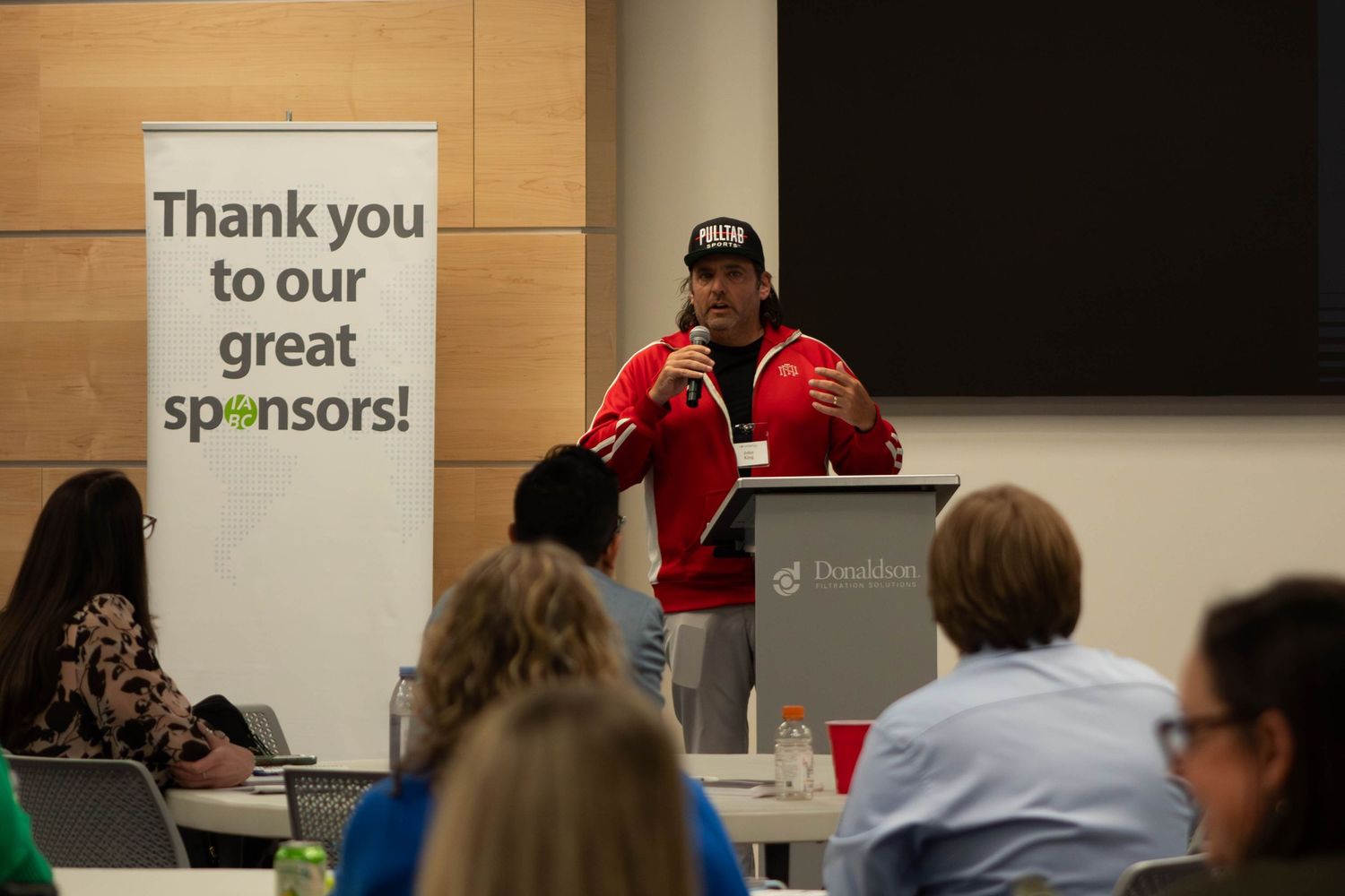 Man in red jacket speaking at podium near Thank You Sponsors banner at a business event.